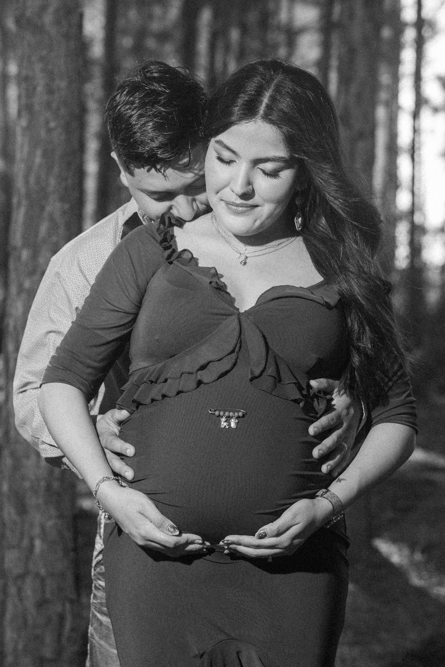 Black and white maternity photo of a couple in a forest, man embracing pregnant woman from behind.
