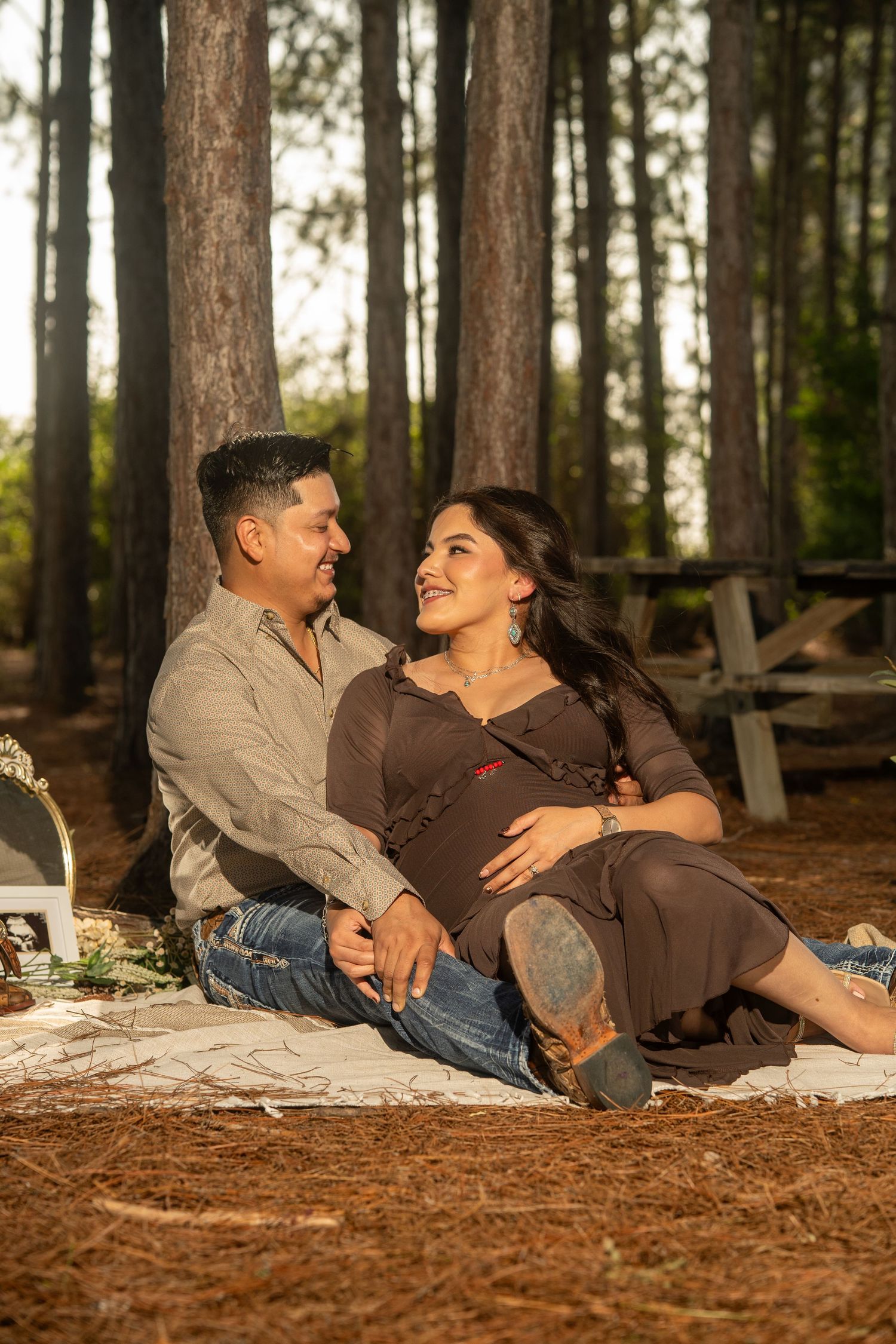 A couple sits on a blanket in a pine forest, smiling at each other during a maternity photo shoot.