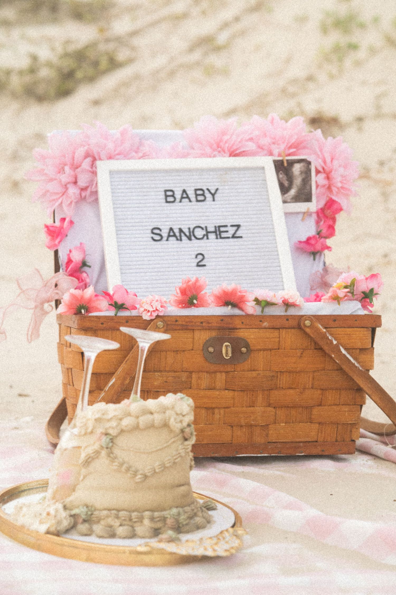 Baby Sanchez 2 pregnancy announcement letterboard with pink flowers on a wicker picnic basket at the beach.