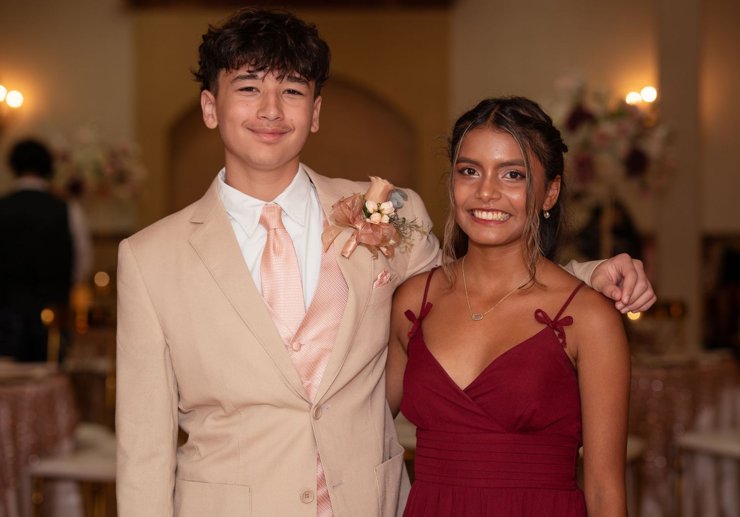 Teen boy in beige suit with boutonniere posing with smiling girl in burgundy gown at a quinceañera.