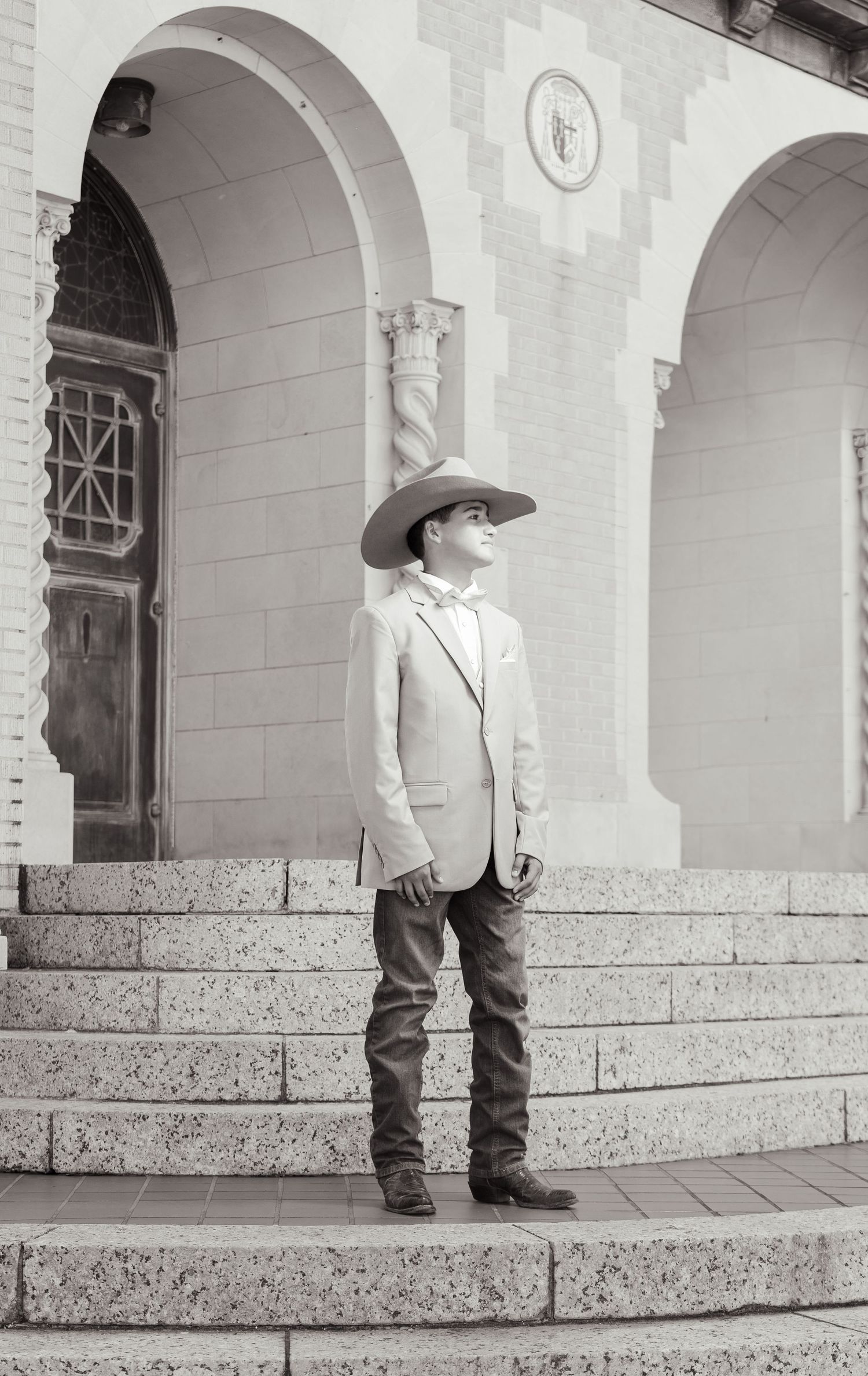 A person in cowboy attire and white blazer stands on stone steps of an arched building entrance in black and white.