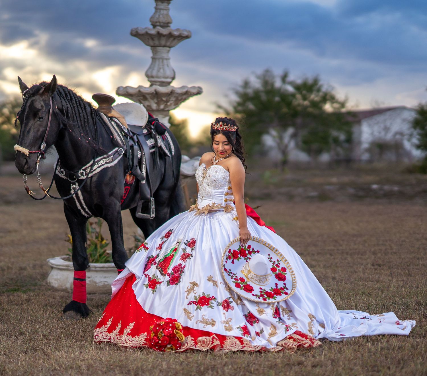 An elegant white ball gown with red floral details photographed outdoors with a horse and decorative fountain.
