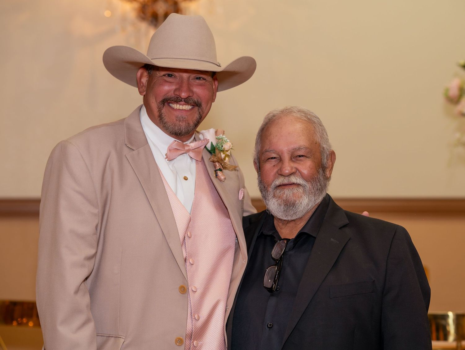 Two men in formal attire pose together, one wearing a cowboy hat and pink suit at a celebration.