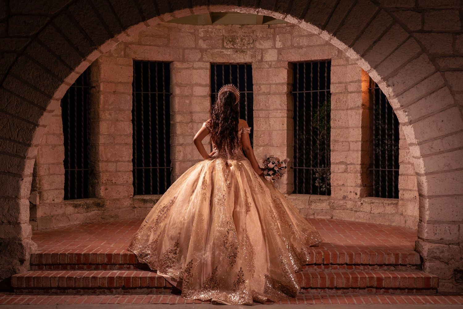 A golden ball gown dress glimmers in the light as a figure stands in an arched stone hallway with barred windows.