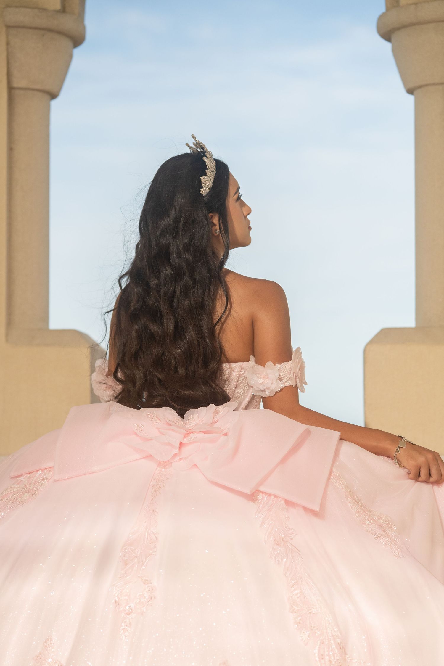 Back view of quinceañera girl in pink off-shoulder gown with bow and hair accessory between columns.