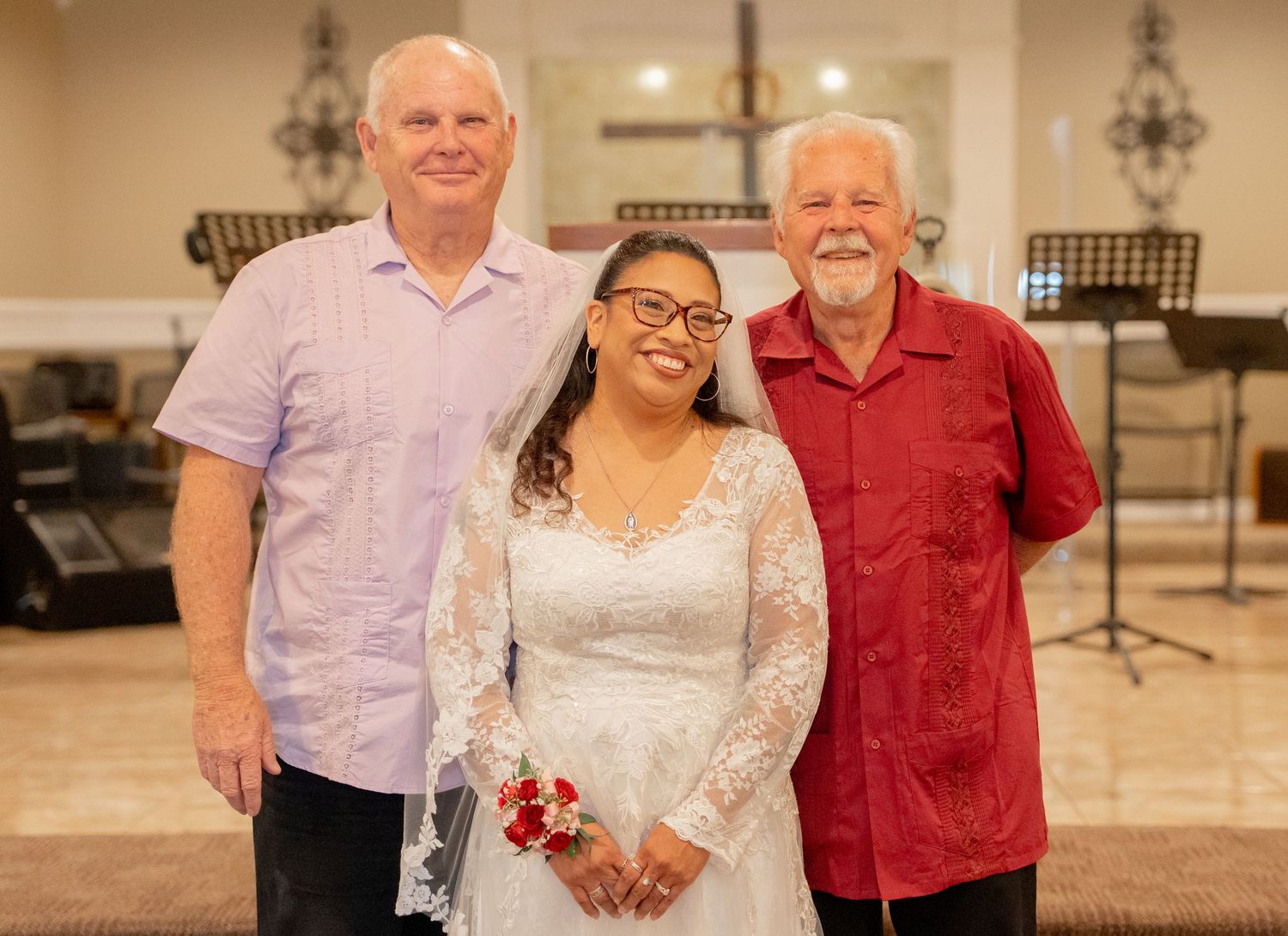 Bride in white lace gown with two older gentlemen posing together inside a church after wedding ceremony.