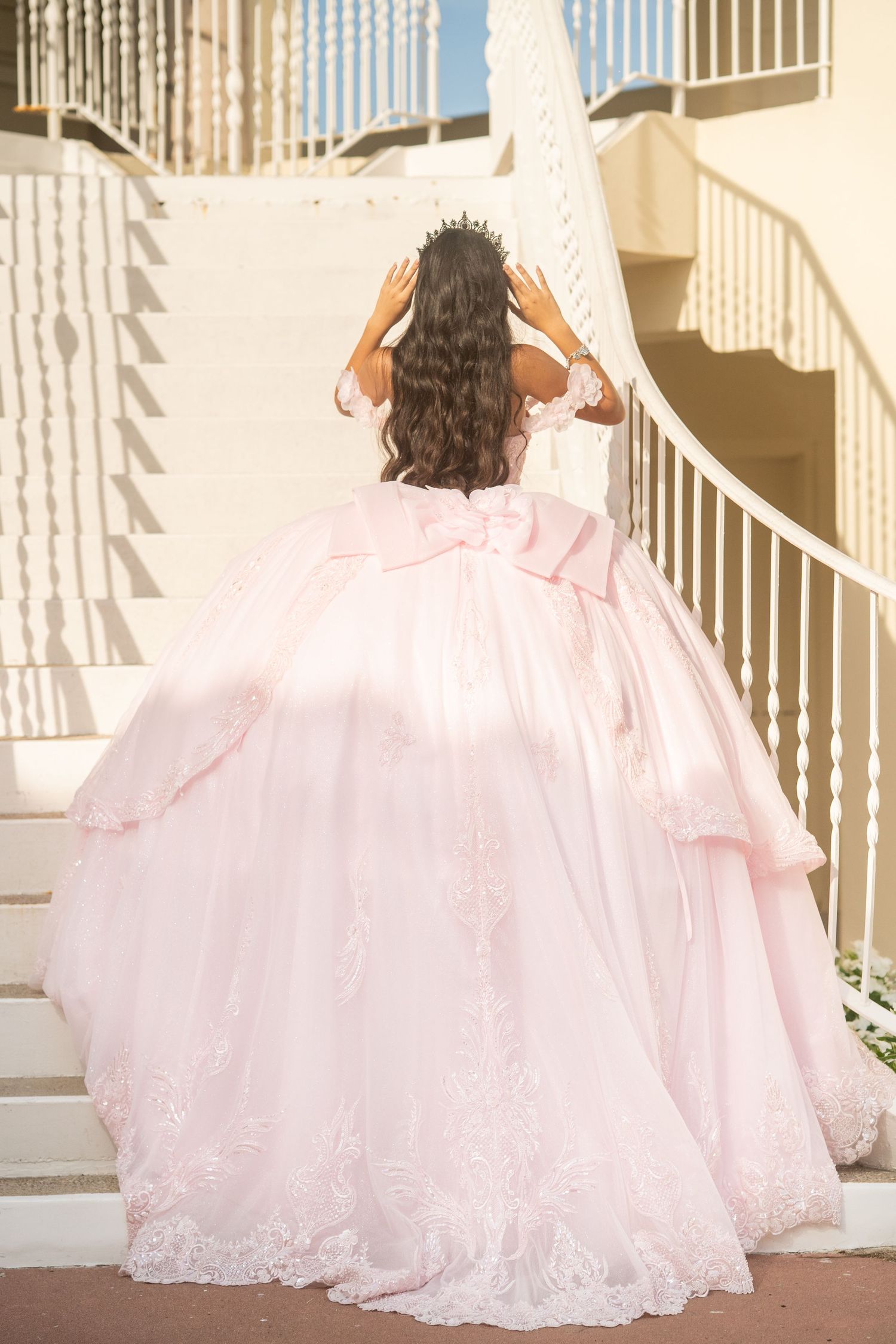 Young woman in elegant pink ball gown with large bow and lace details ascending white staircase, wearing a tiara.