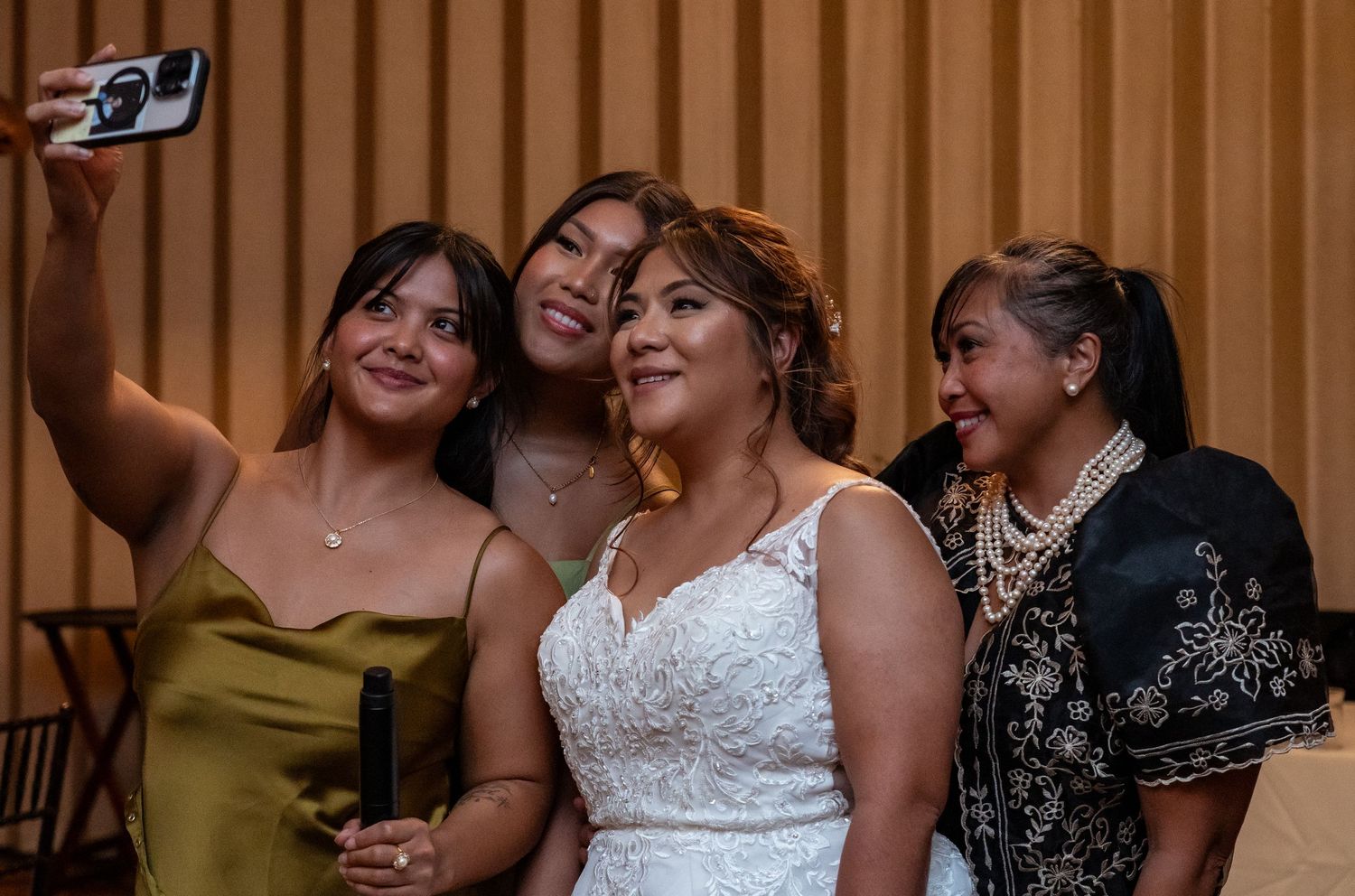 Bride in white lace gown takes selfie with three friends at wedding reception.