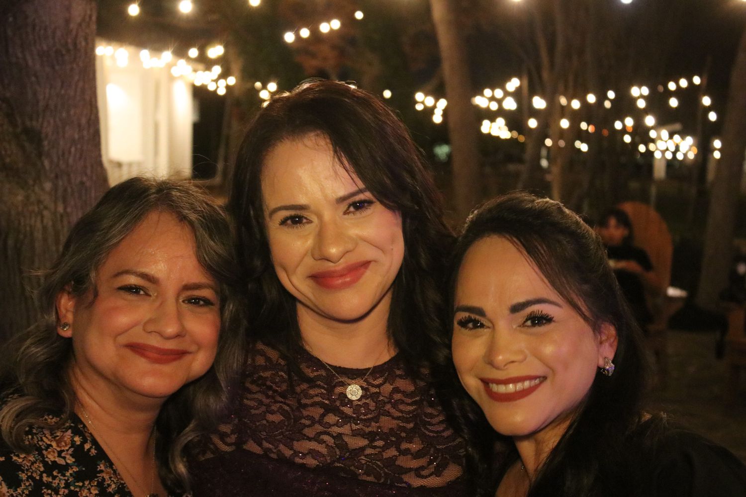 Three women smiling at an evening outdoor event with string lights twinkling in the background.