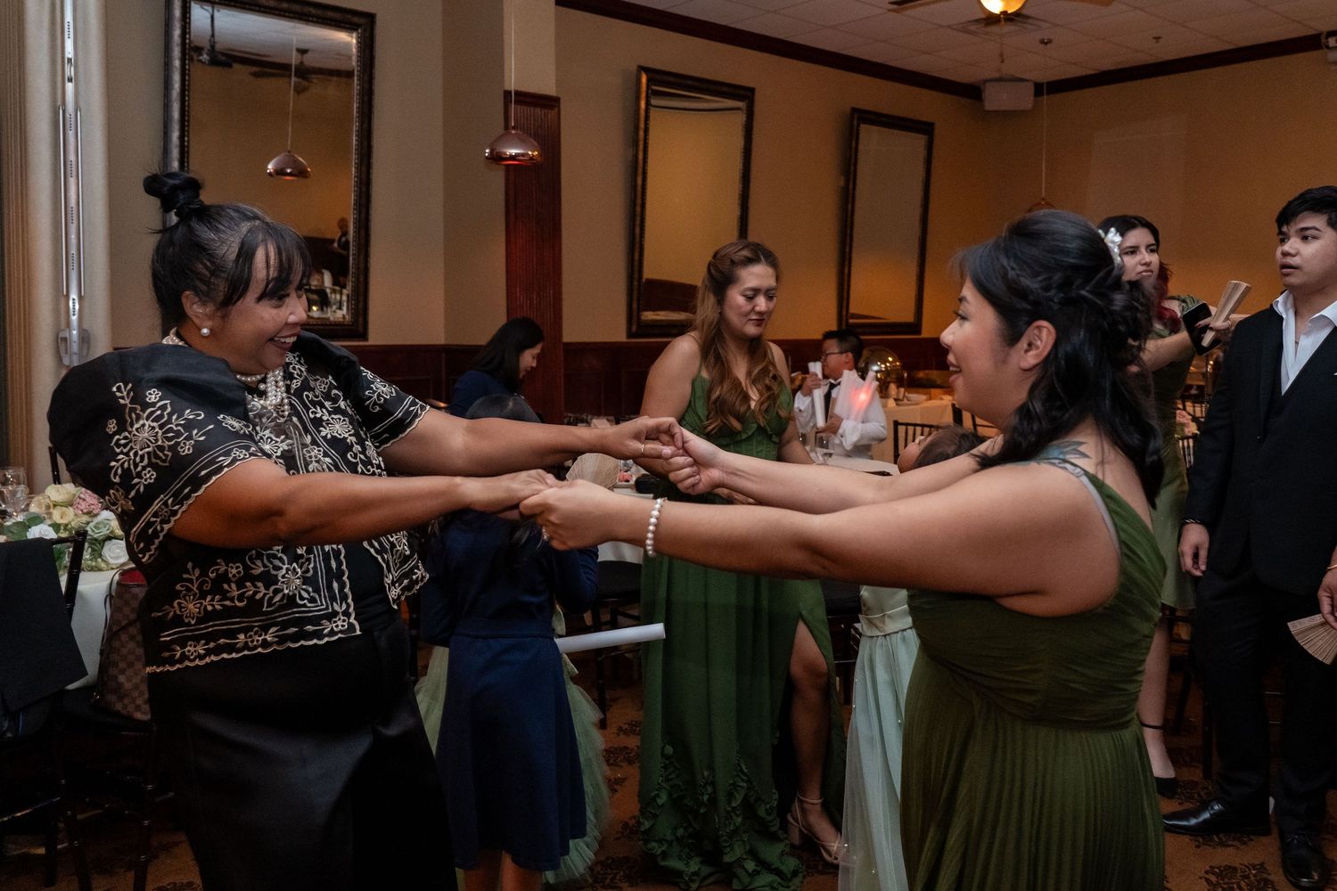 Two women hold hands dancing at a formal event, with guests in elegant attire seated in the background.