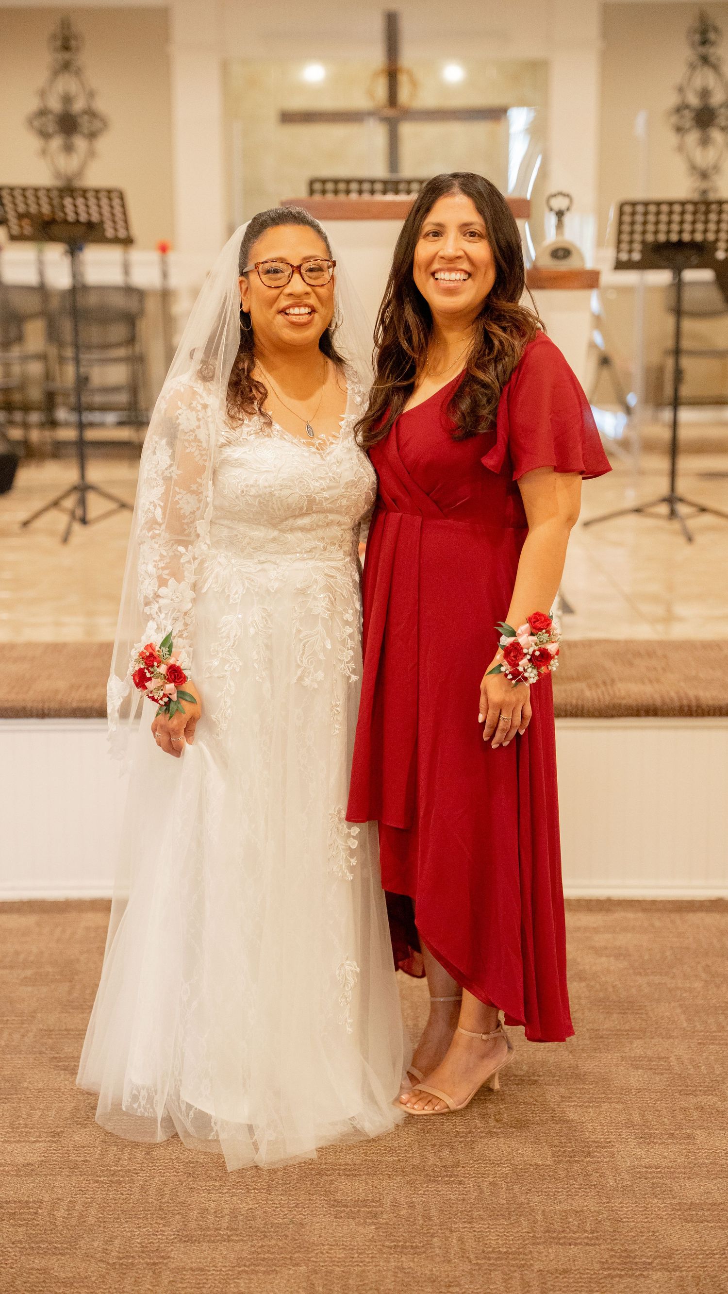Bride in lace gown poses with bridesmaid in red dress at church wedding reception.