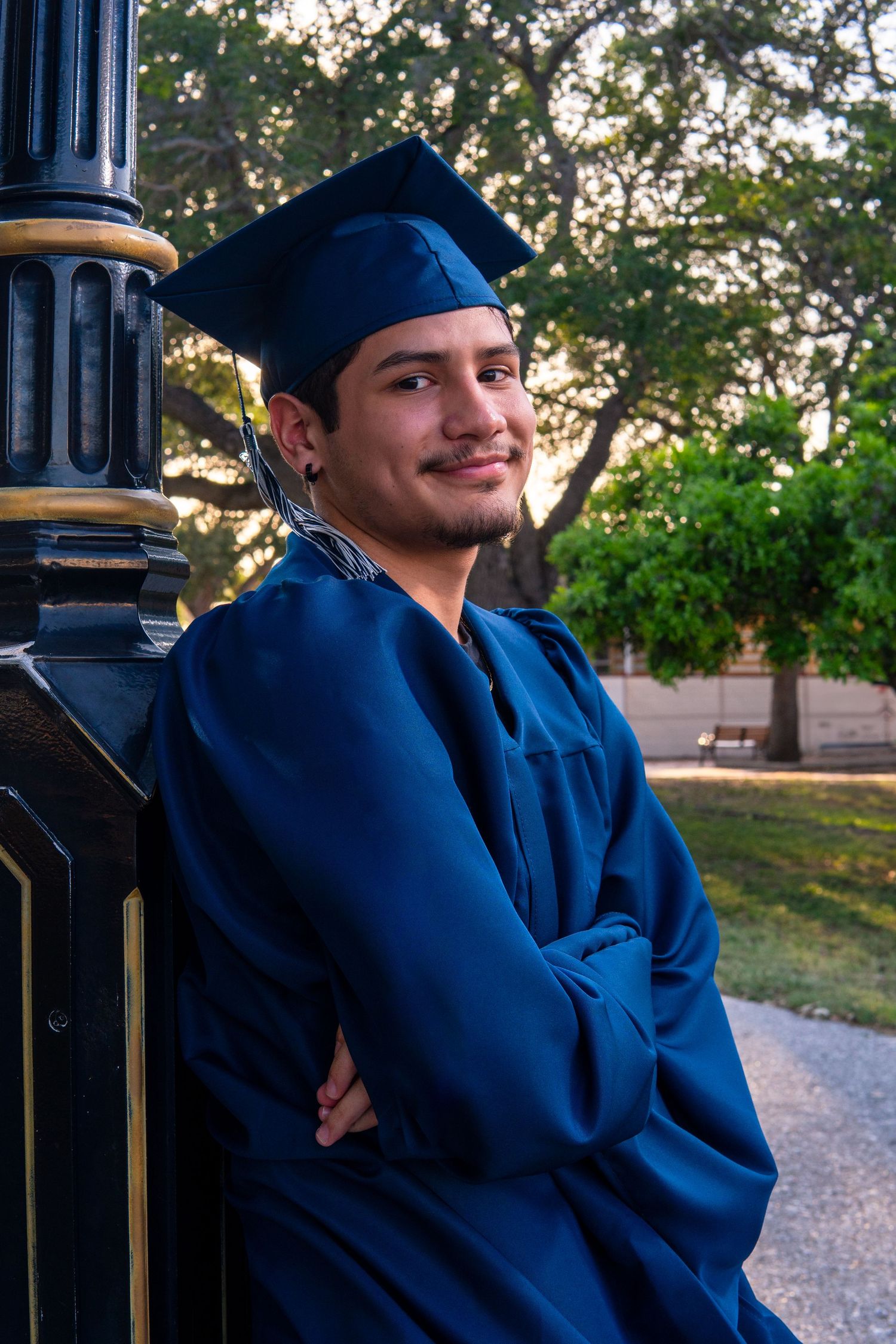 A graduate in dark blue cap and gown leans against a decorative lamp post at dusk during commencement.