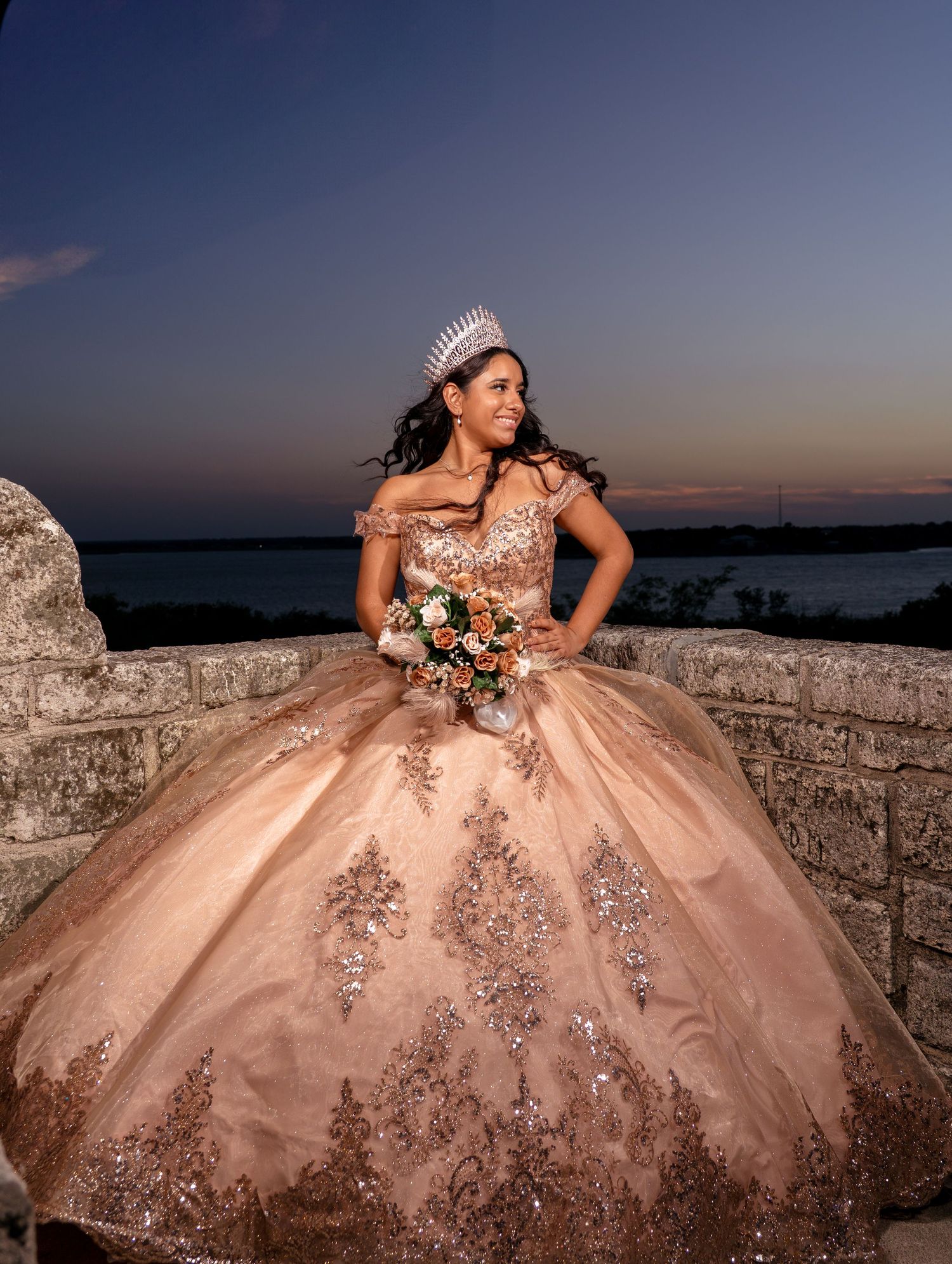 Sunset photoshoot of a quinceañera wearing a sparkling rose gold ballgown posed along stone walls overlooking the ocean.
