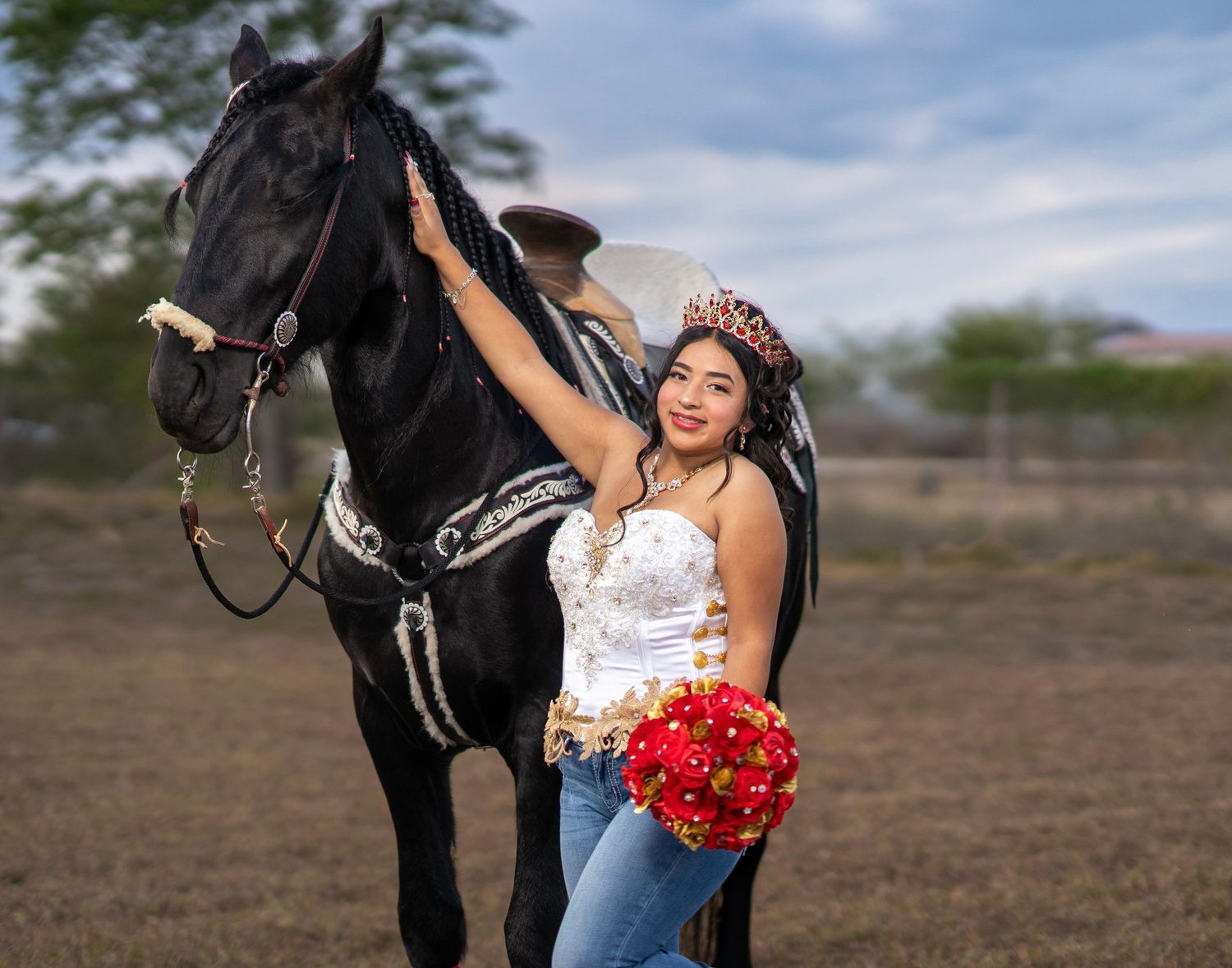 A woman in jeans poses with a black horse and red bouquet in an outdoor field setting.