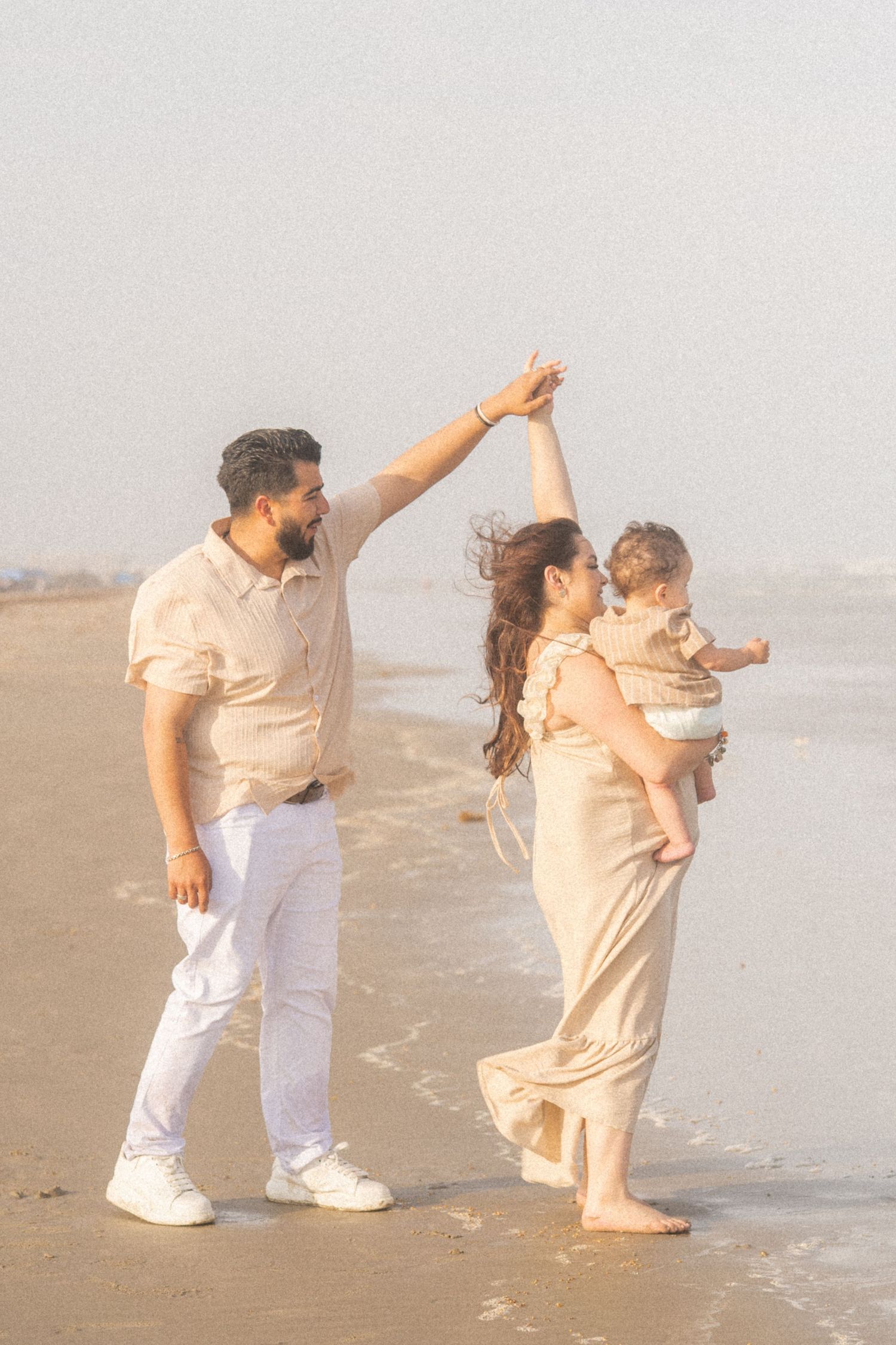 A joyful family of three enjoys a beach day, with dad twirling mom who holds their baby on the shoreline.