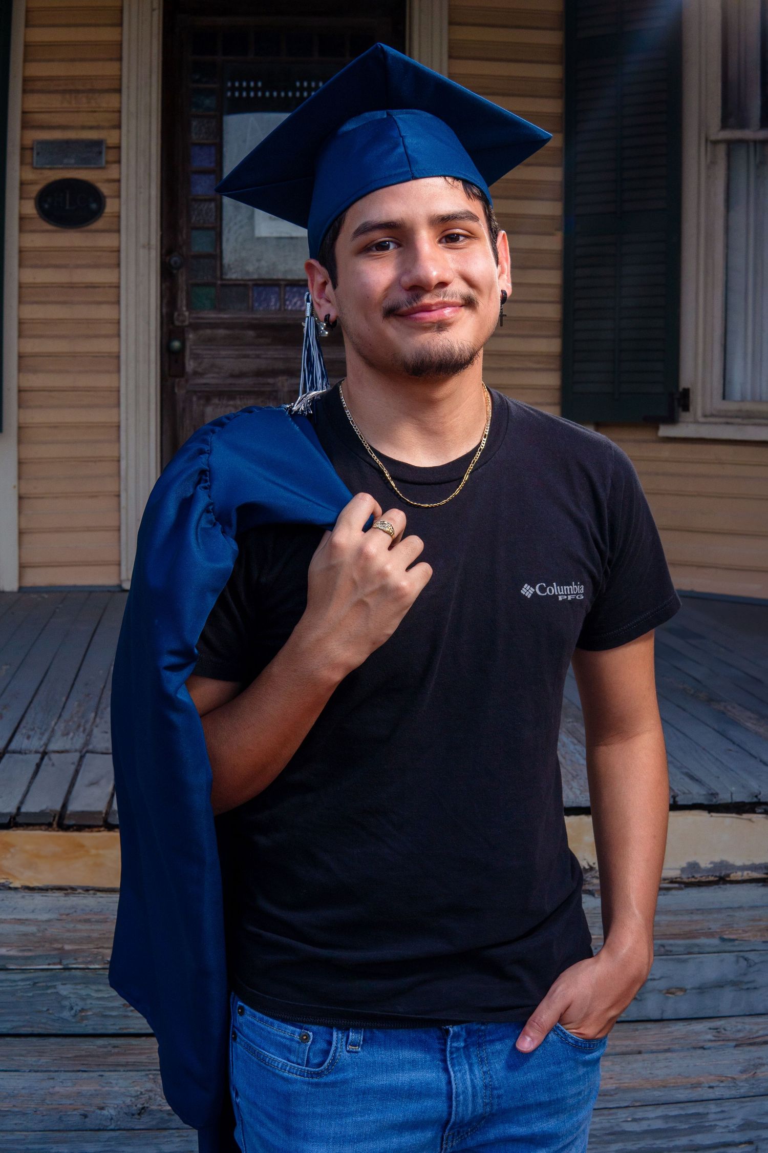 A graduate in a navy blue cap and gown stands on a porch wearing a black Columbia t-shirt and jeans.