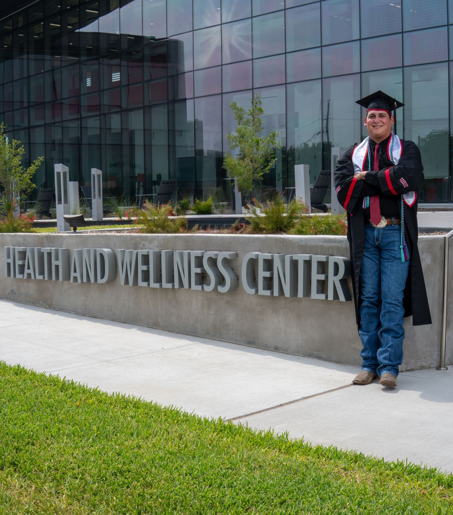 A graduate in cap and gown stands by the entrance sign of a modern Health and Wellness Center building.