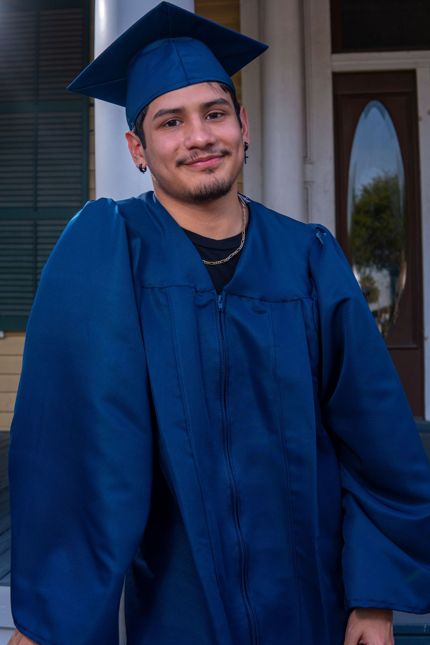 Graduate wearing blue cap and gown stands smiling on graduation day in front of white building.