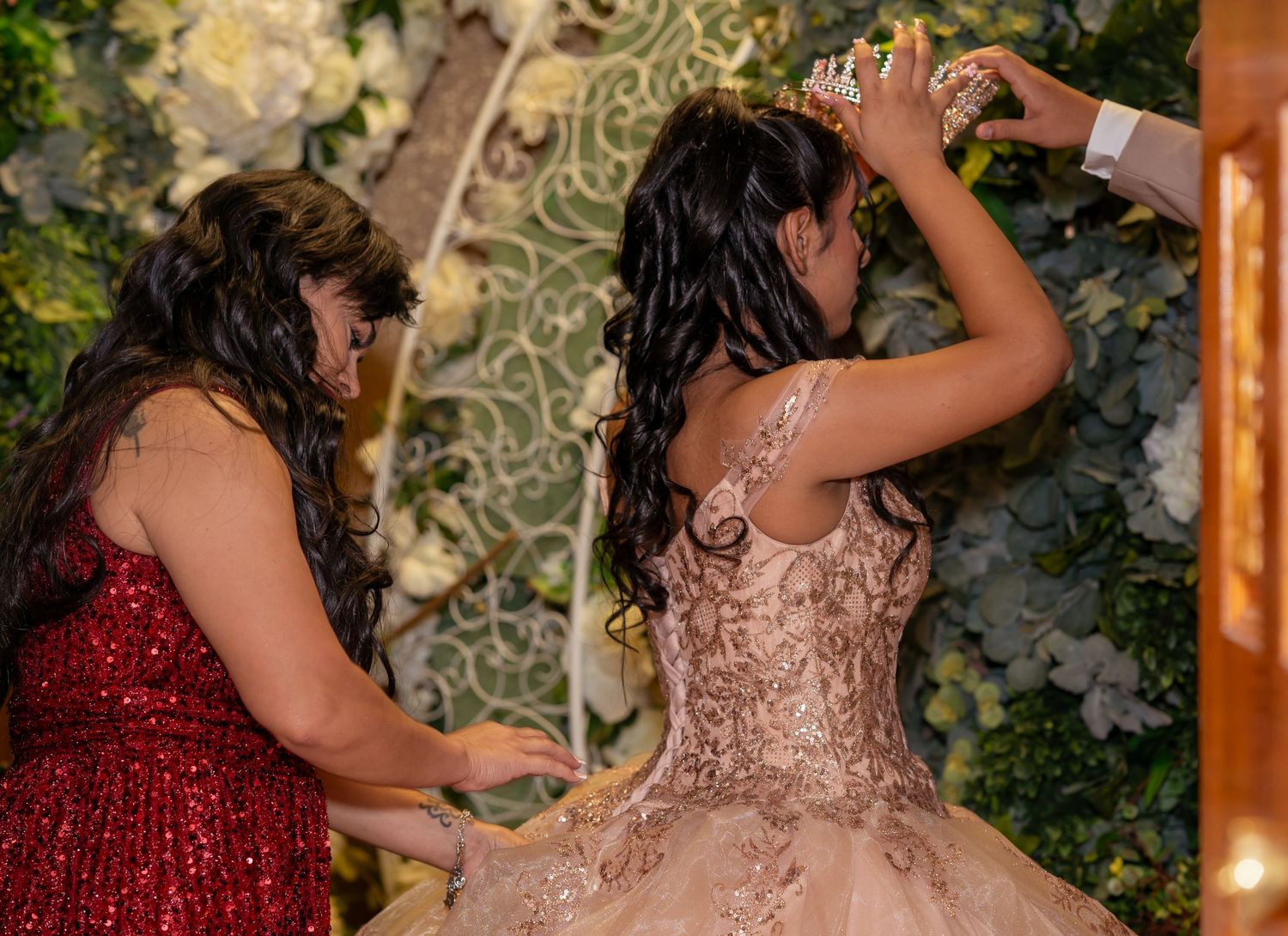 Two women in formal gowns at a quinceañera celebration, one in red sequins helping adjust a crown on the other.