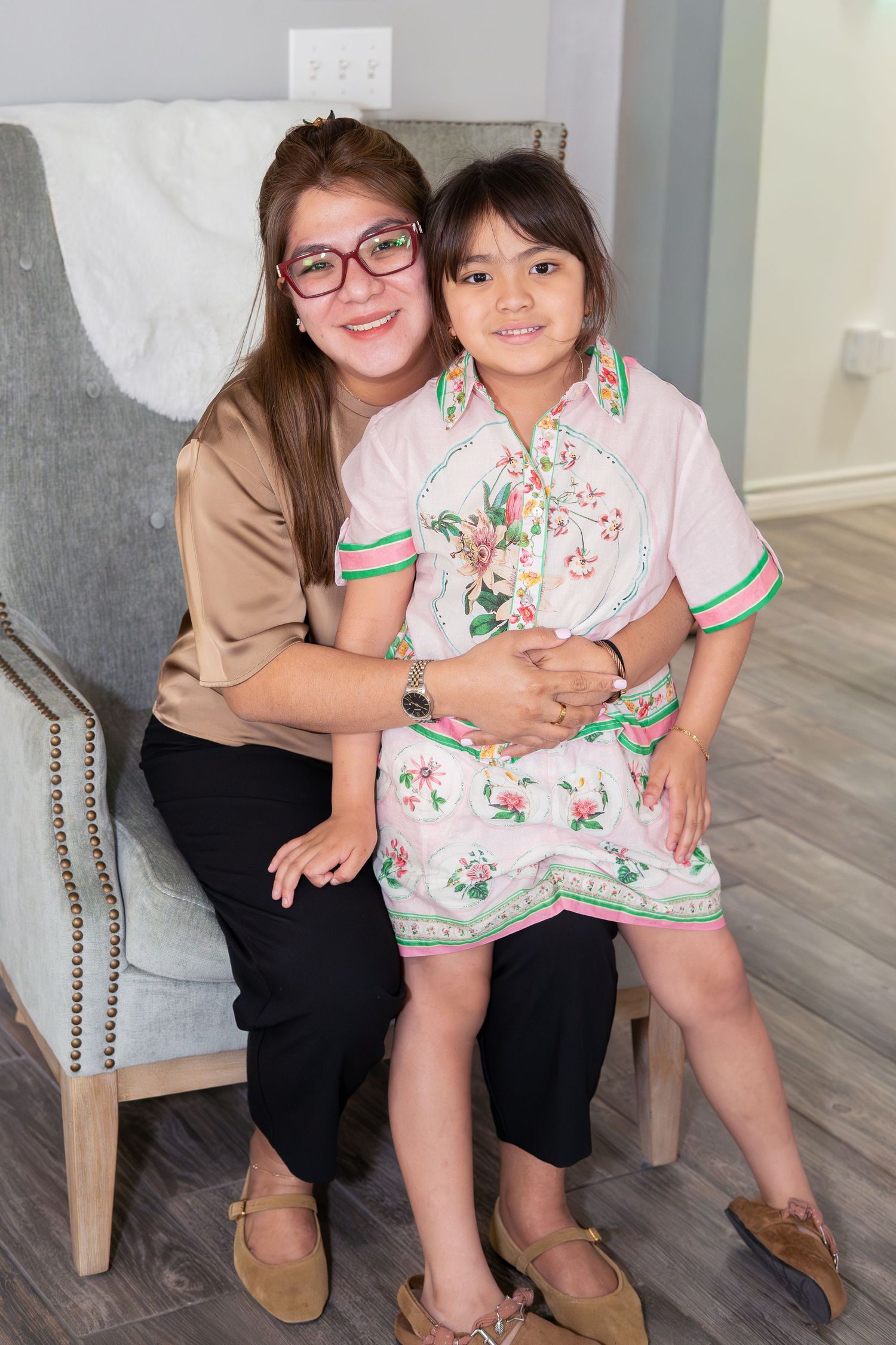 Woman with glasses hugging young girl in floral dress while seated on chair, smiling warmly.