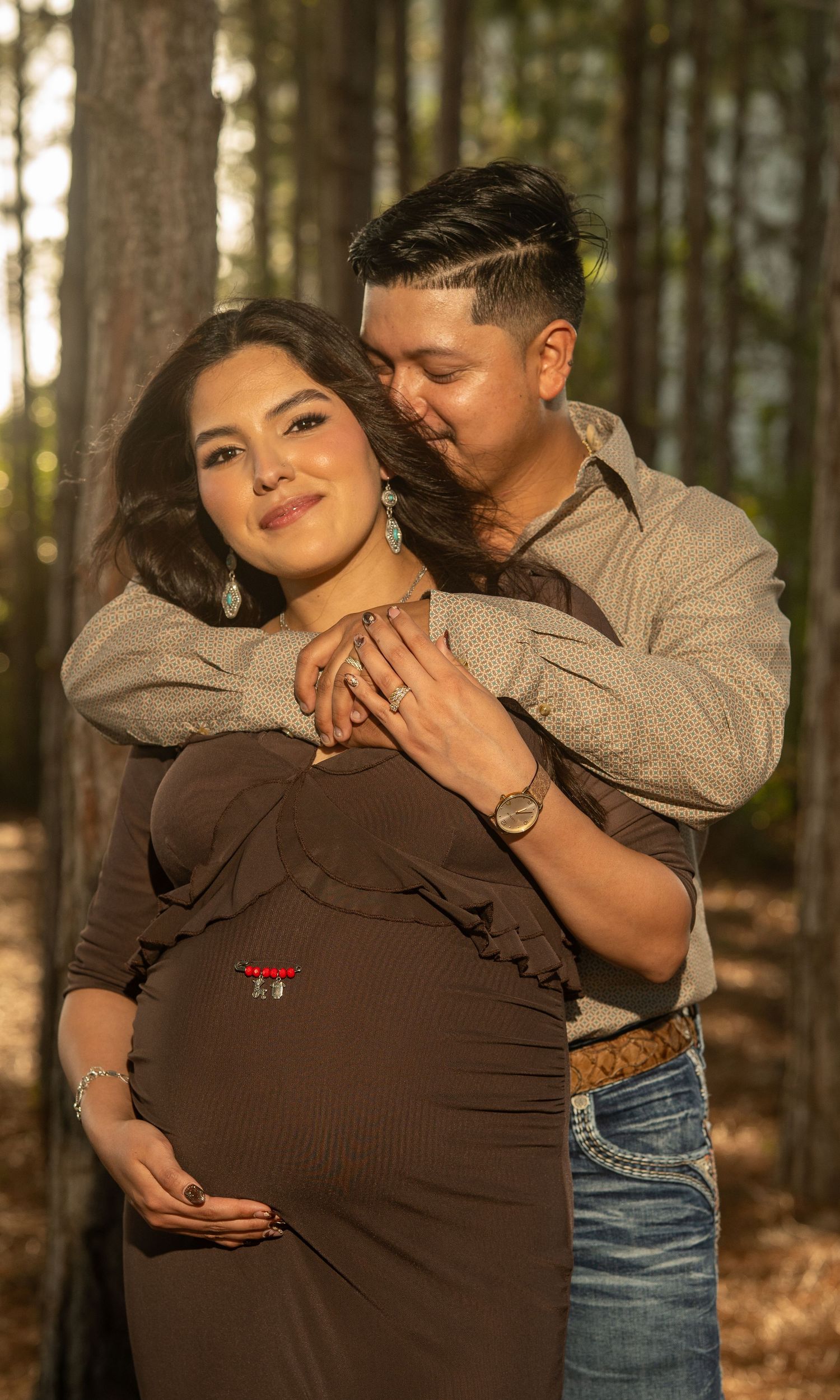 Couple maternity photo in forest, woman in brown dress cradling baby bump while partner embraces her from behind.