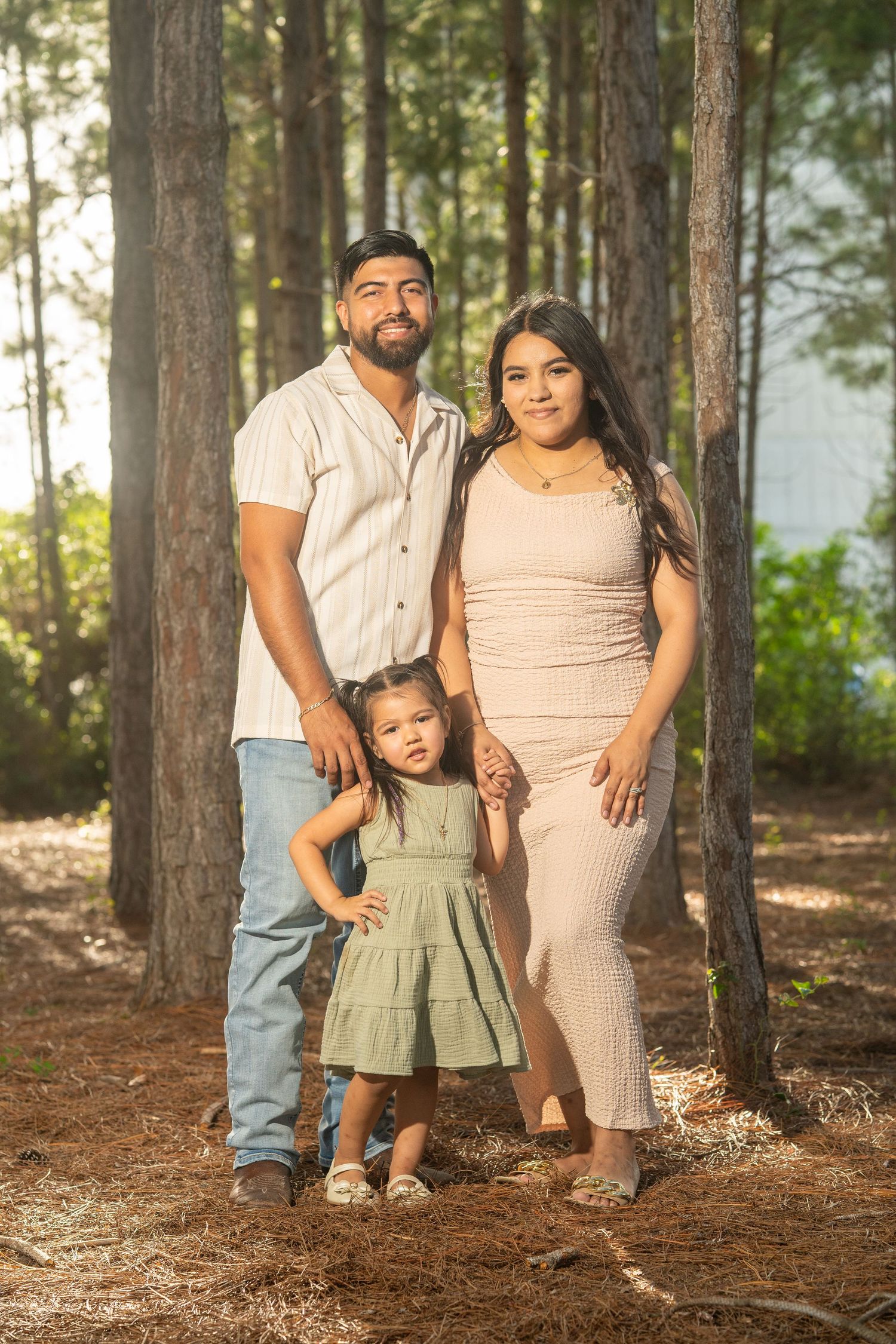 A young Hispanic family of three poses among pine trees, with parents flanking their small daughter in a green dress.