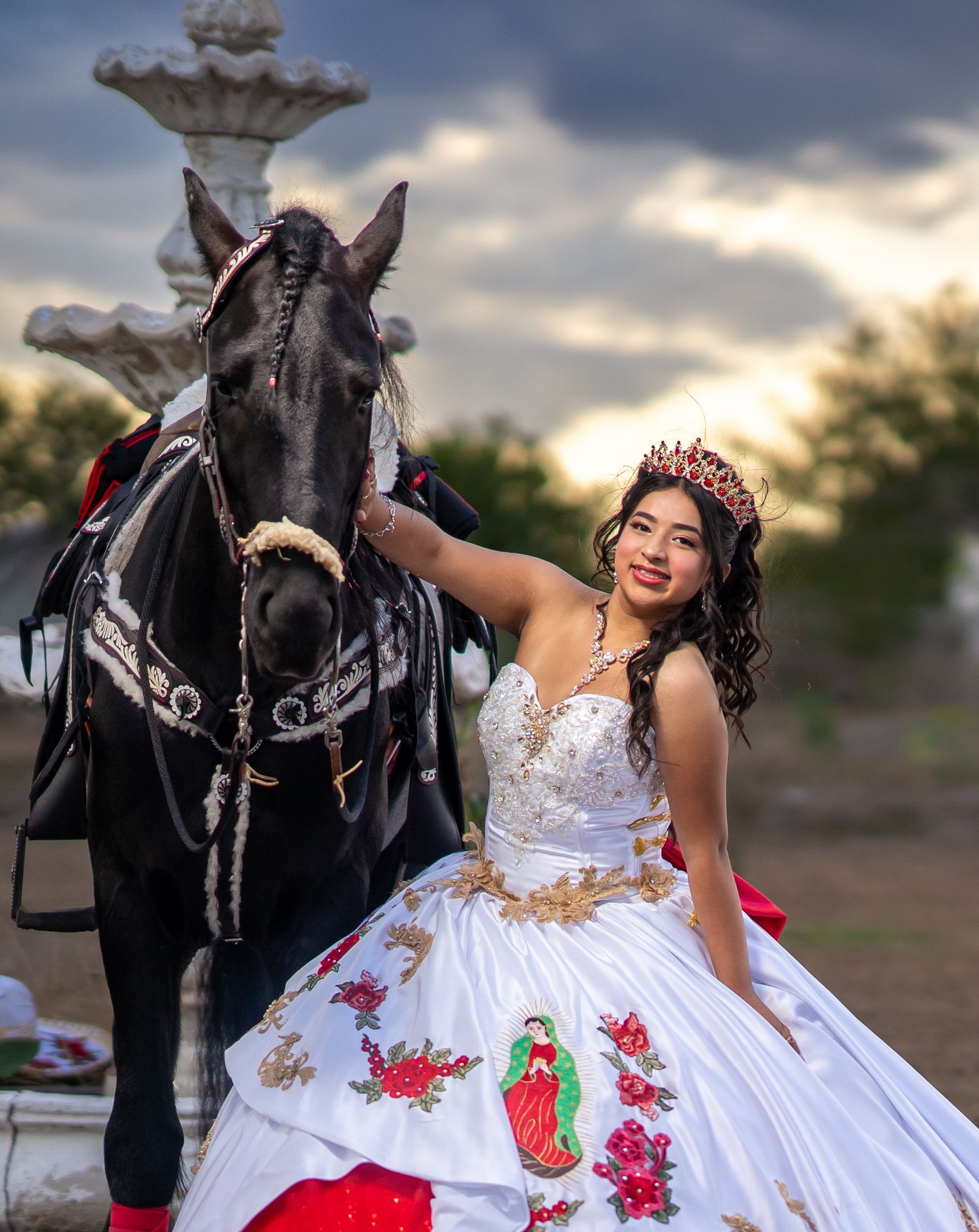 A white and red embroidered dress flows elegantly while posing with a black horse against a sunset sky.