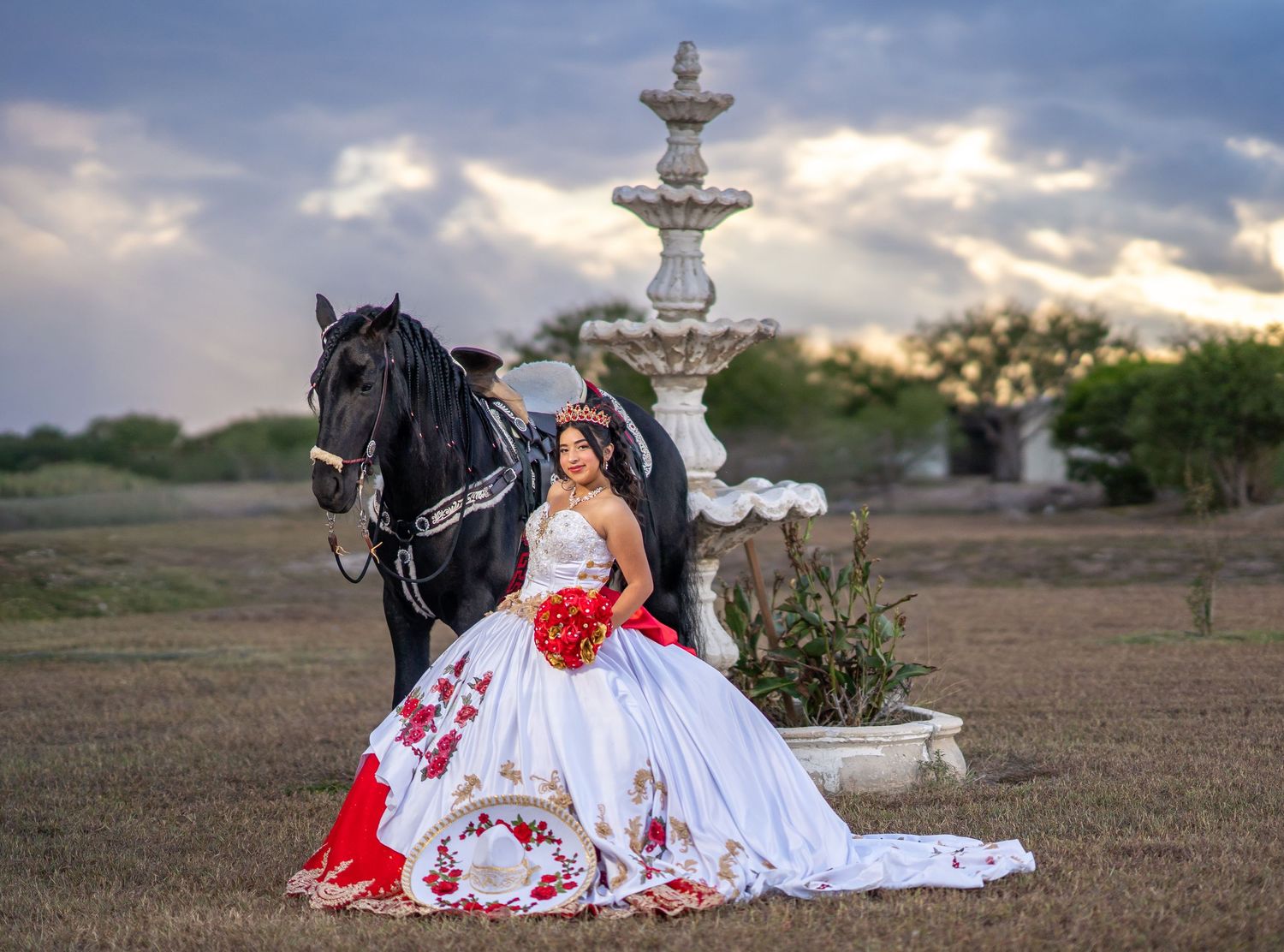 A white dress with red accents flows across a desert landscape as a black horse stands nearby at sunset.