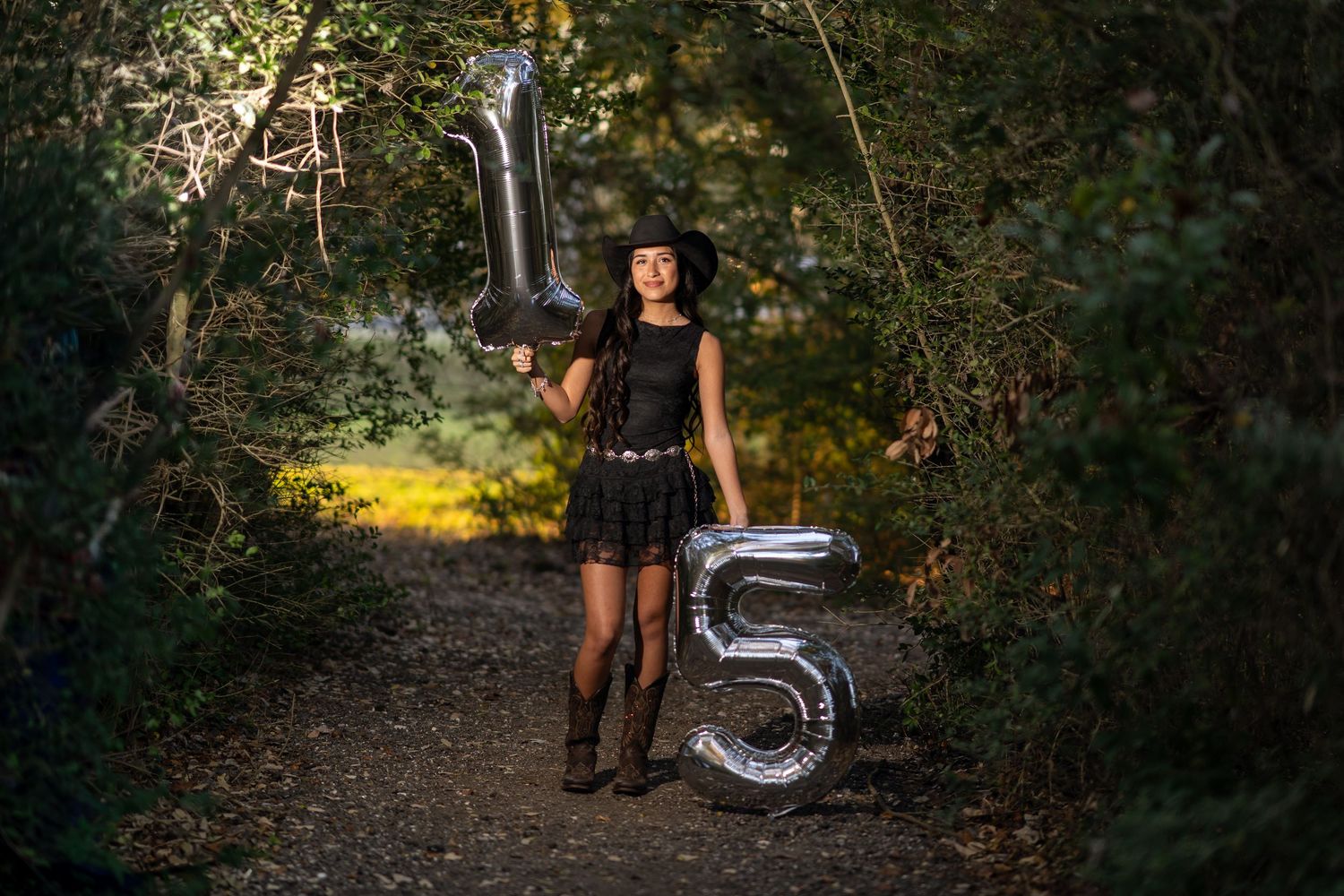 Teen girl in black dress and hat holds silver number 15 balloons on a forest path for quinceañera photos.