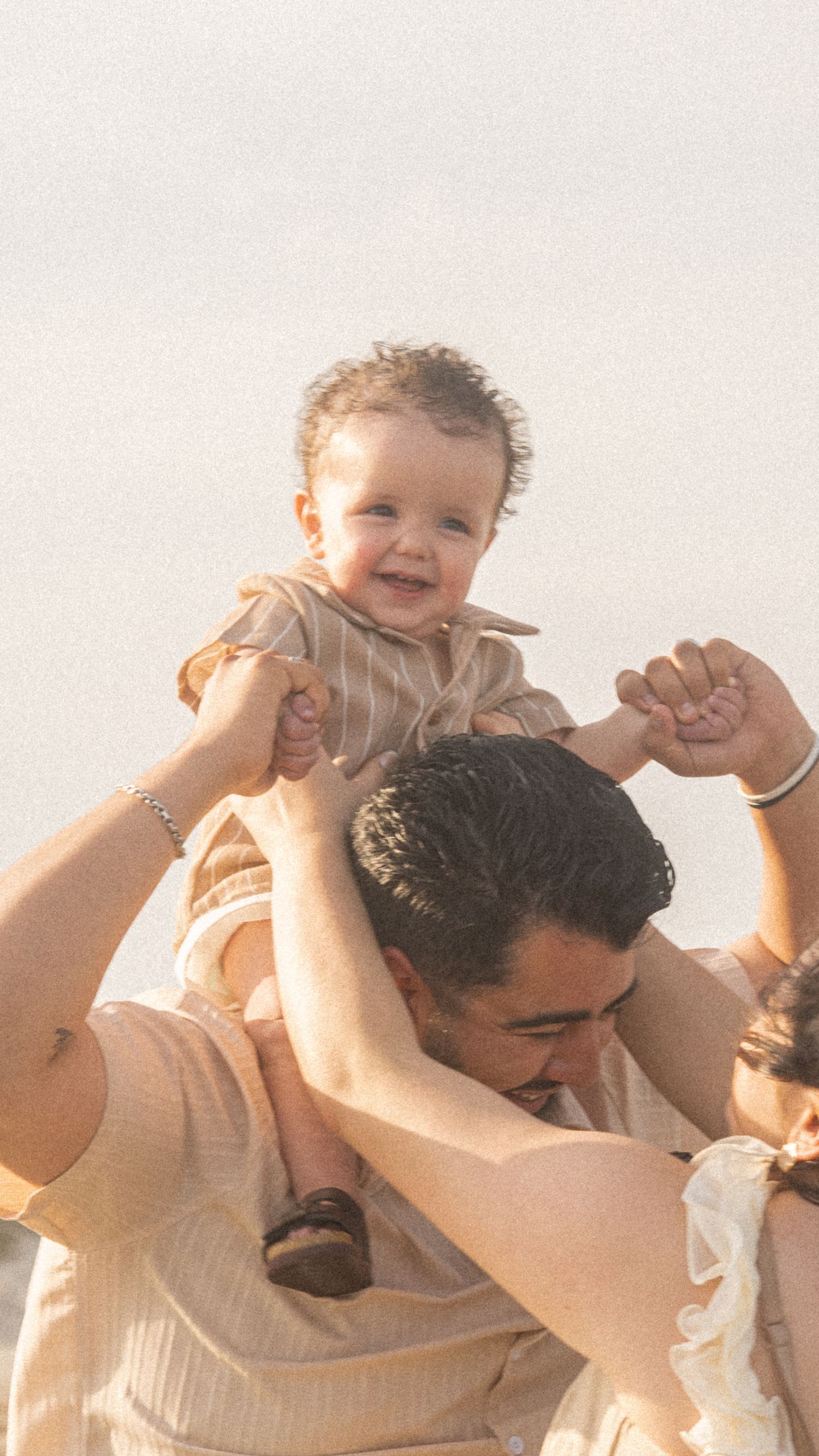 A smiling baby sits on a man's shoulders outdoors, held by two adults in warm tones against a bright sky.
