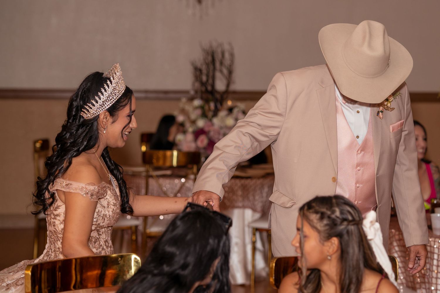 A quinceañera girl in a rose gold gown and tiara interacts with a man in a tan cowboy hat.