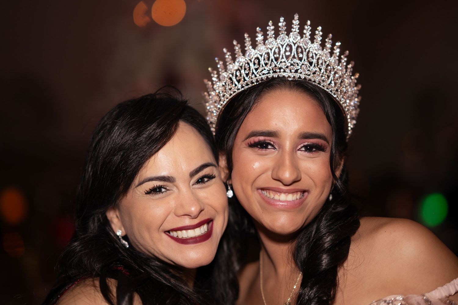Two smiling women pose together at a quinceañera, one wearing an elegant silver crown and off-shoulder gown.