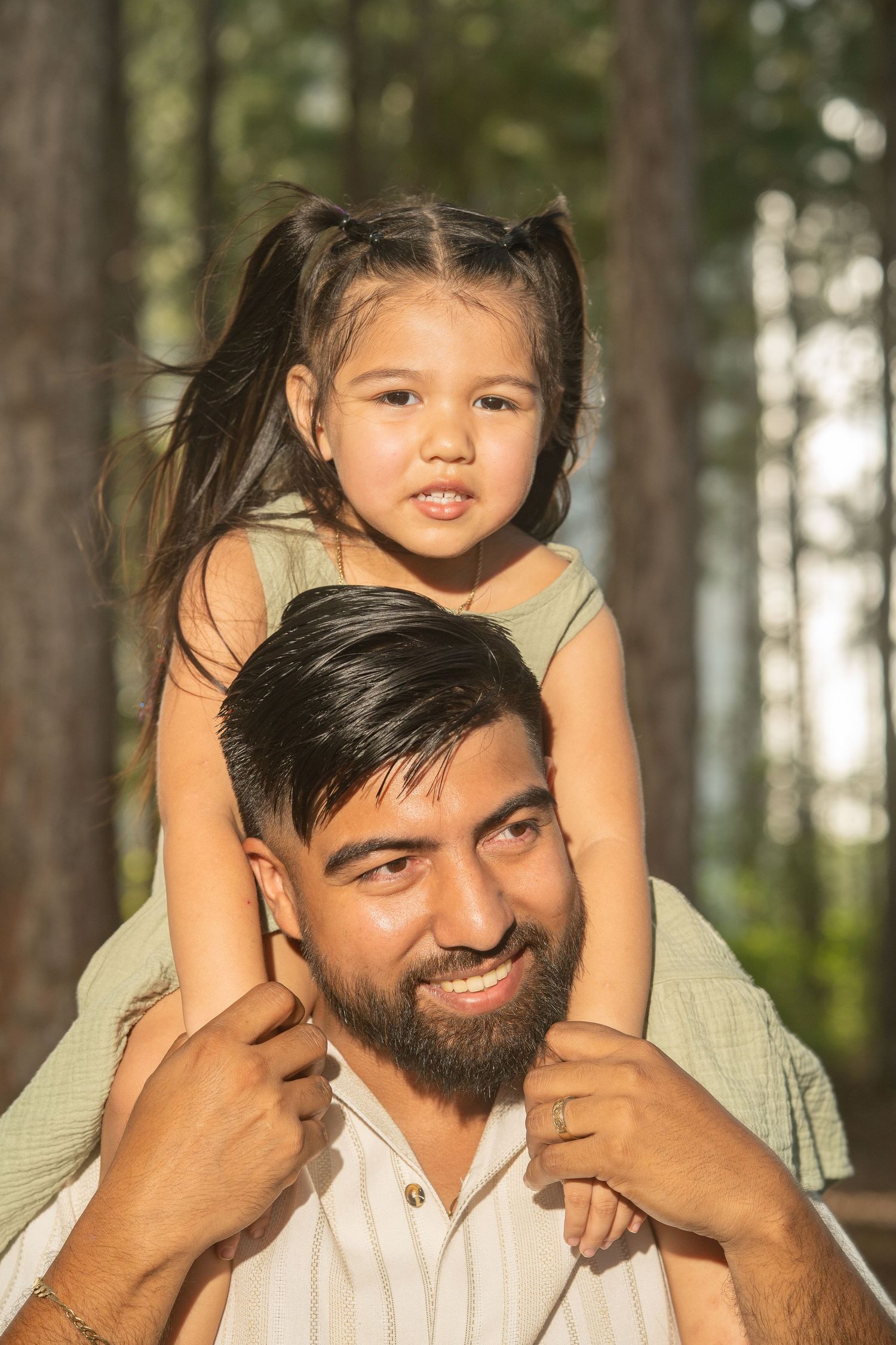 Smiling bearded father carries young daughter on his shoulders in a sunlit forest setting.