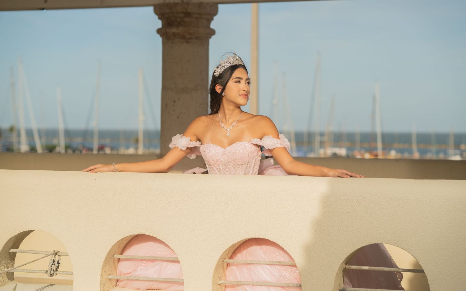 Young woman in pink ball gown and tiara poses gracefully on a balcony overlooking a marina at sunset.