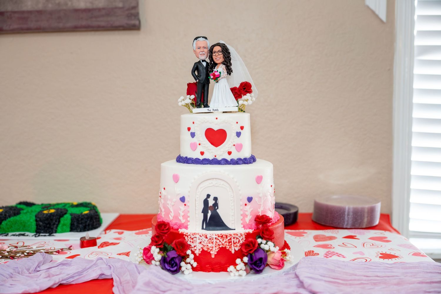 Three-tier white wedding cake with bride and groom topper, heart decorations, and silhouette design on decorated table.