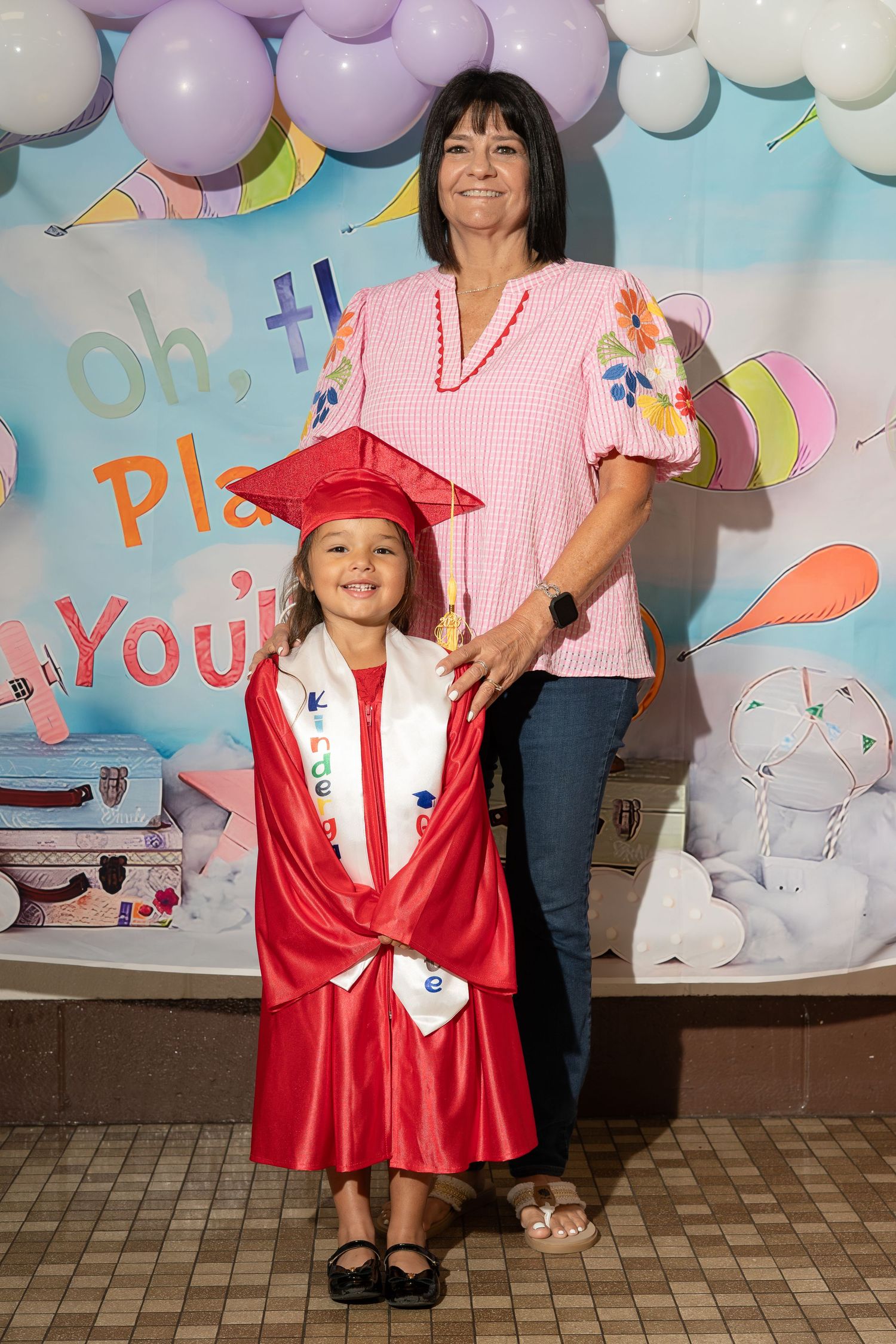 Young girl in red graduation cap and gown poses with woman at kindergarten graduation ceremony.