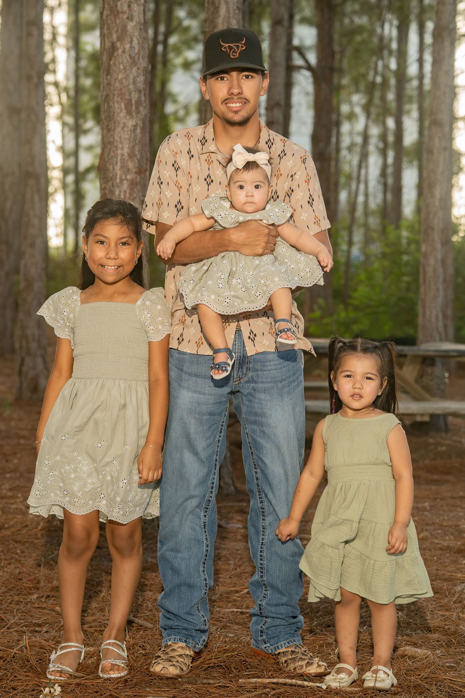 A father holds his baby while standing with two young daughters in matching sage dresses among pine trees outdoors.