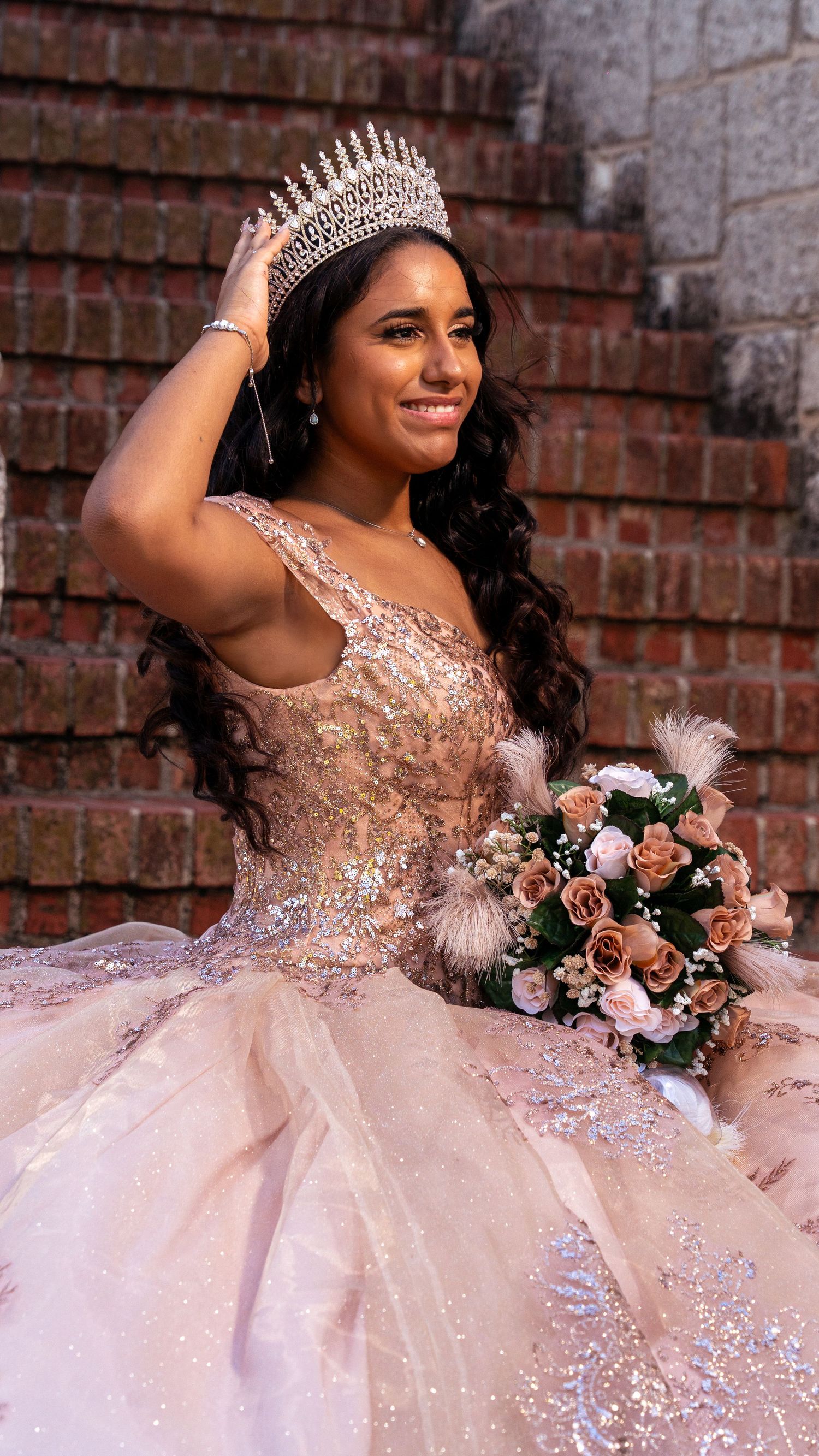 A sparkling pink quinceanera dress with intricate beading and lace details against a brick wall backdrop.