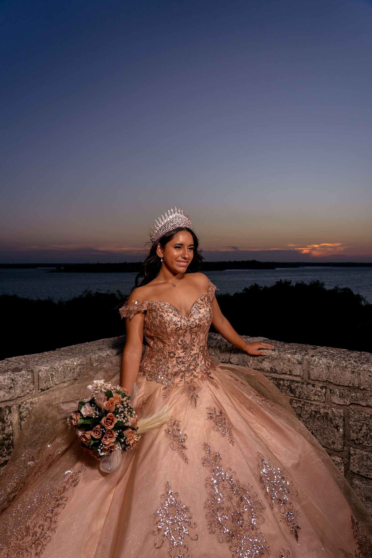 A stunning rose gold ball gown photographed at sunset against a water view.