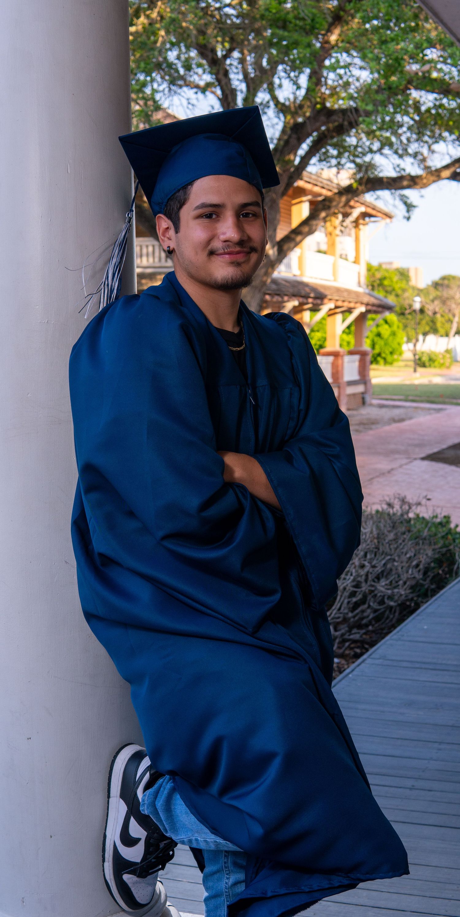 Graduate in blue cap and gown poses confidently against a white column on a sunny outdoor campus.