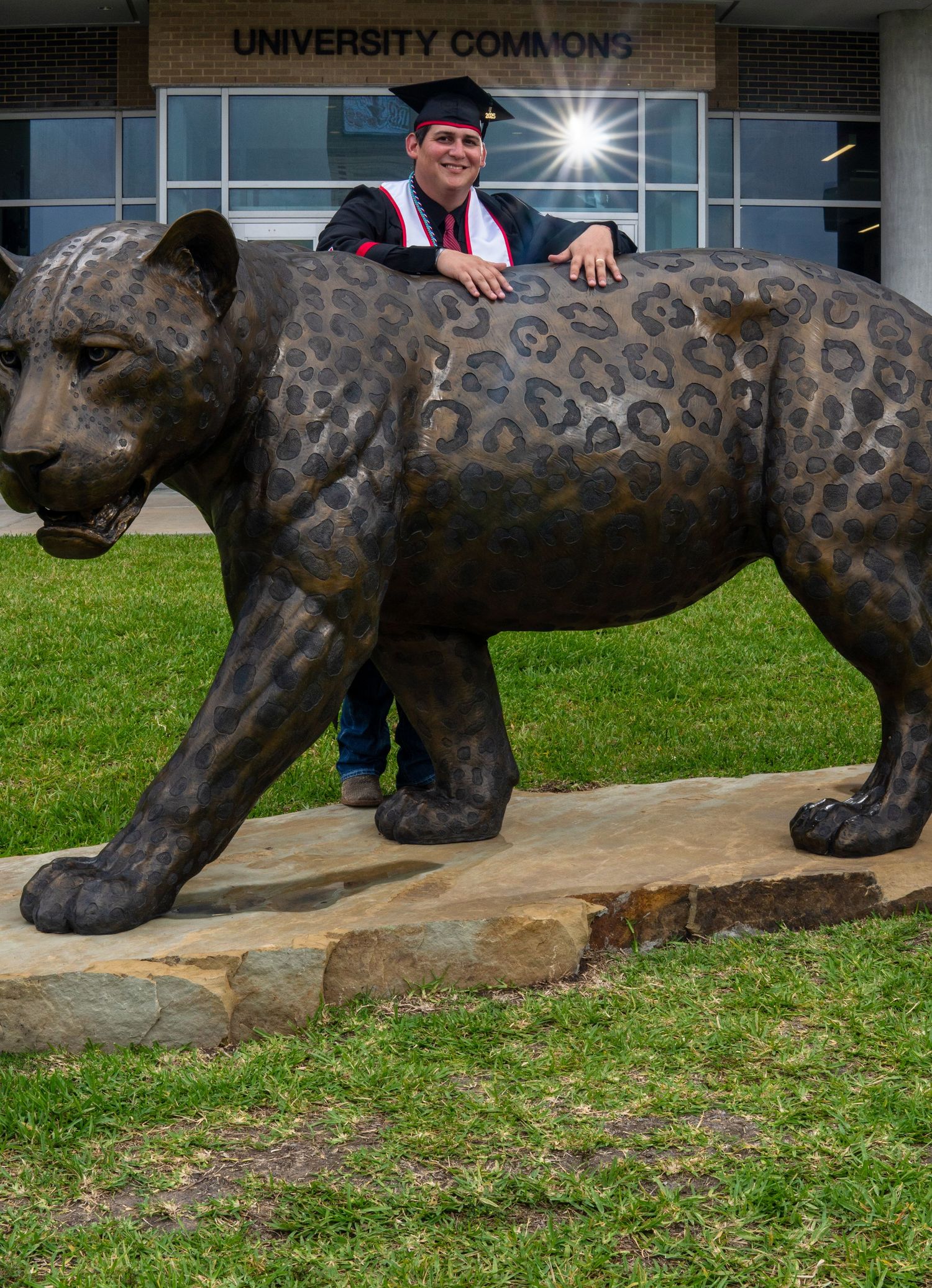 Graduate in cap and gown poses with large bronze jaguar statue outside University Commons building.