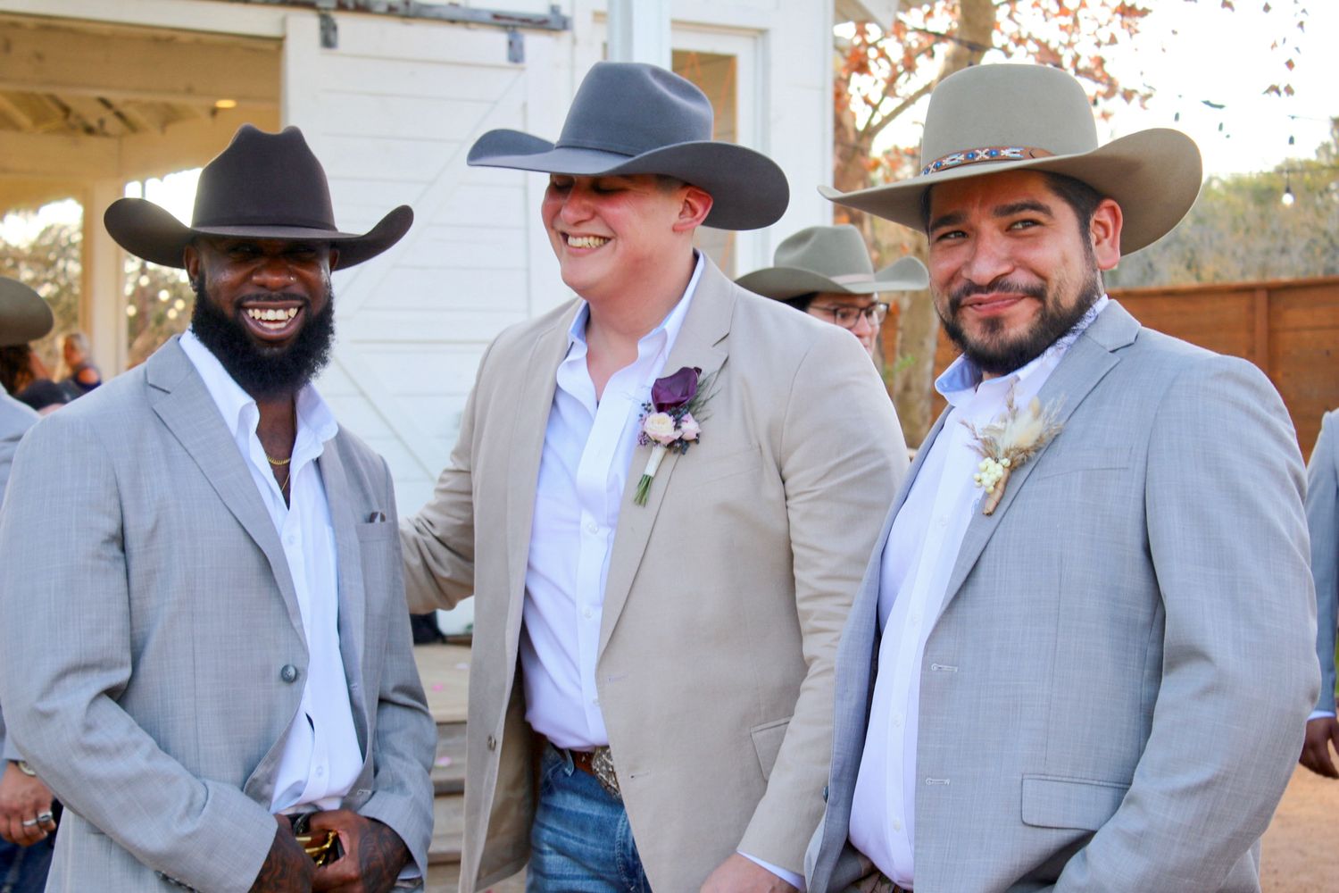 Three wedding guests in matching light gray suits, white shirts and cowboy hats smile together at outdoor venue.