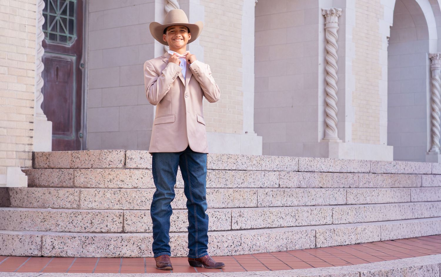 A person wearing a cowboy hat and light-colored jacket poses on stone steps with arched architecture in the background.