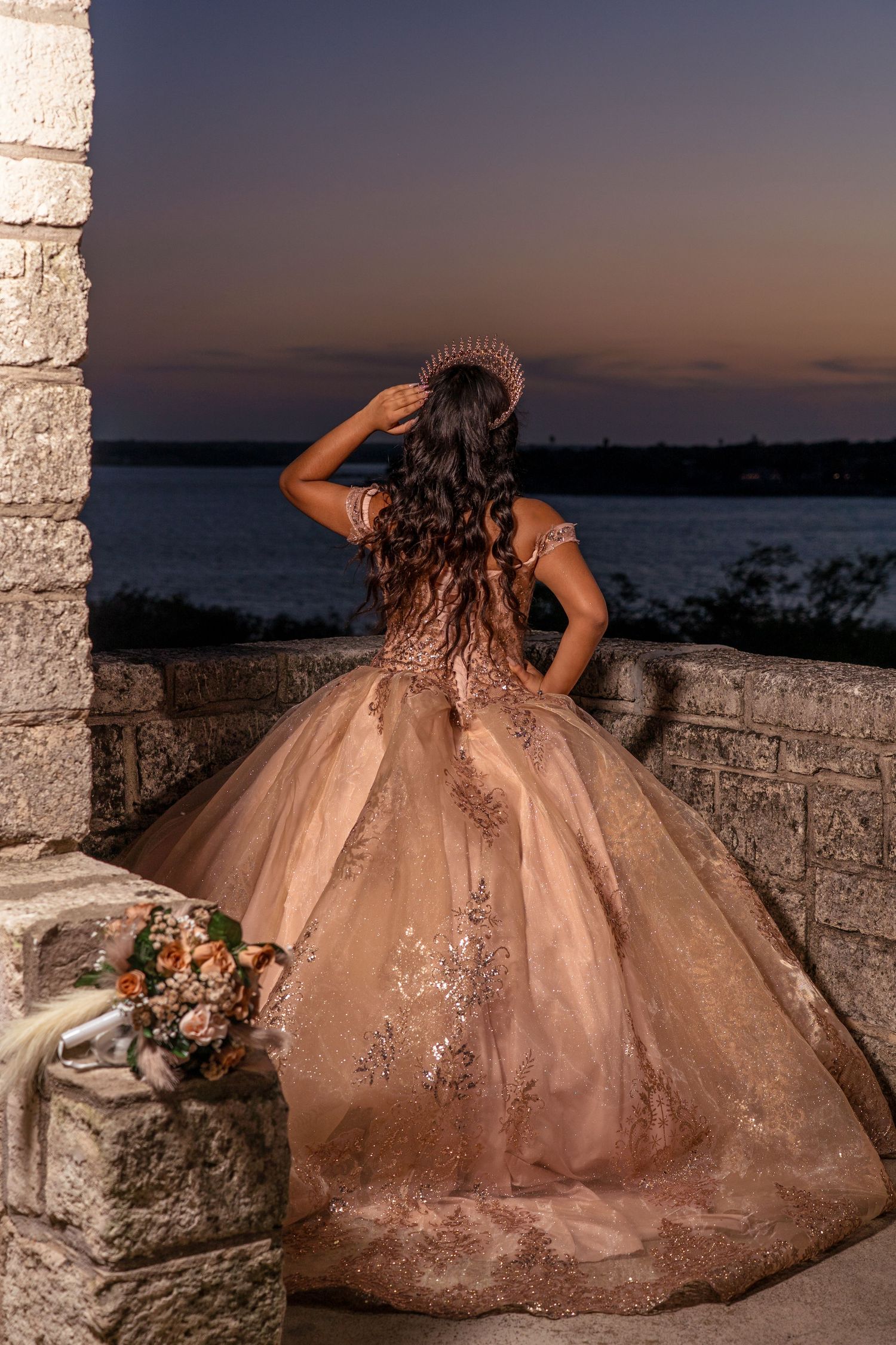 Ornate blush ballgown with lace details photographed at dusk against a stone balcony overlooking the ocean.