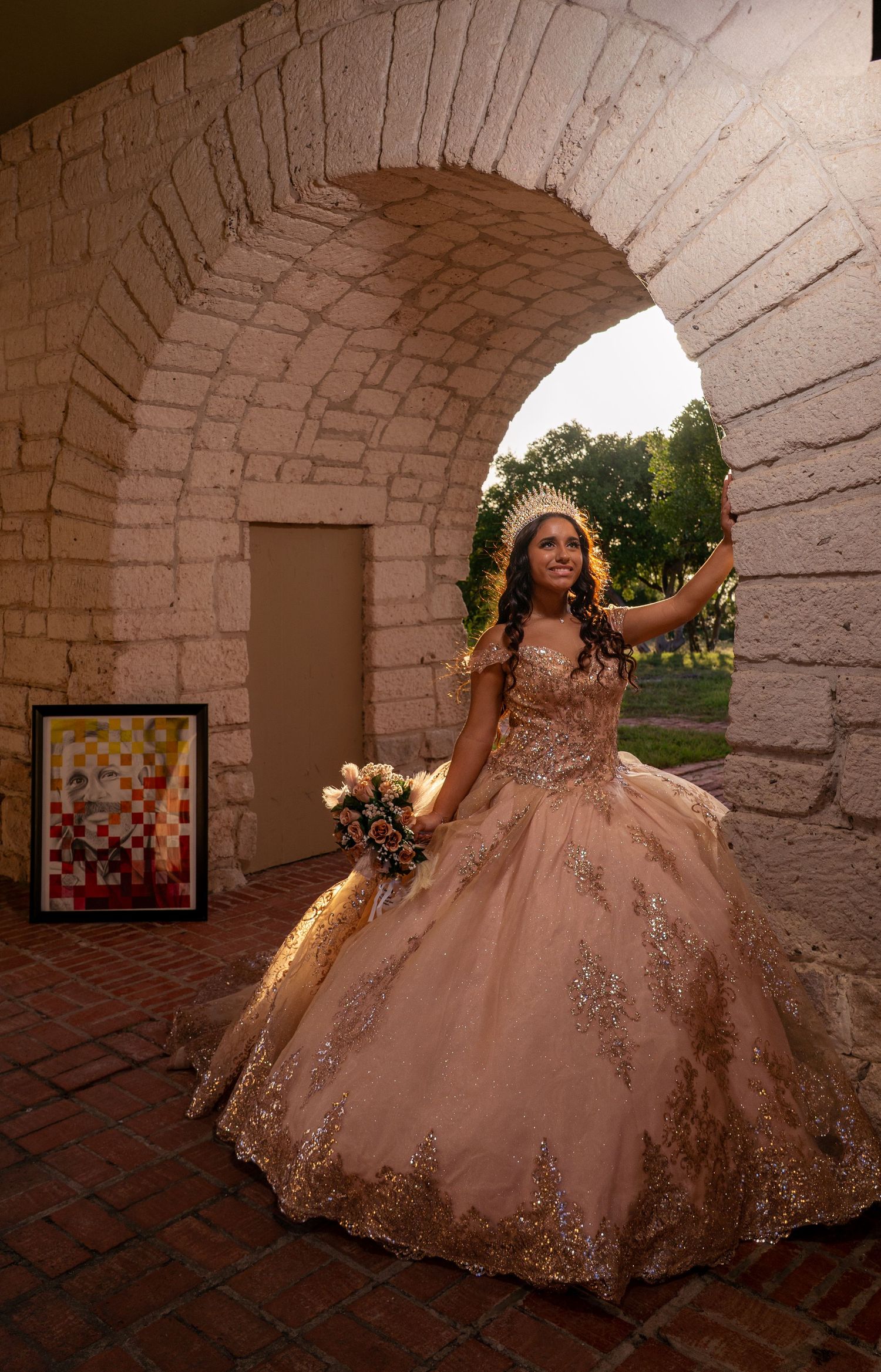 A person in an ornate rose gold quinceanera gown poses beneath a stone archway at sunset.