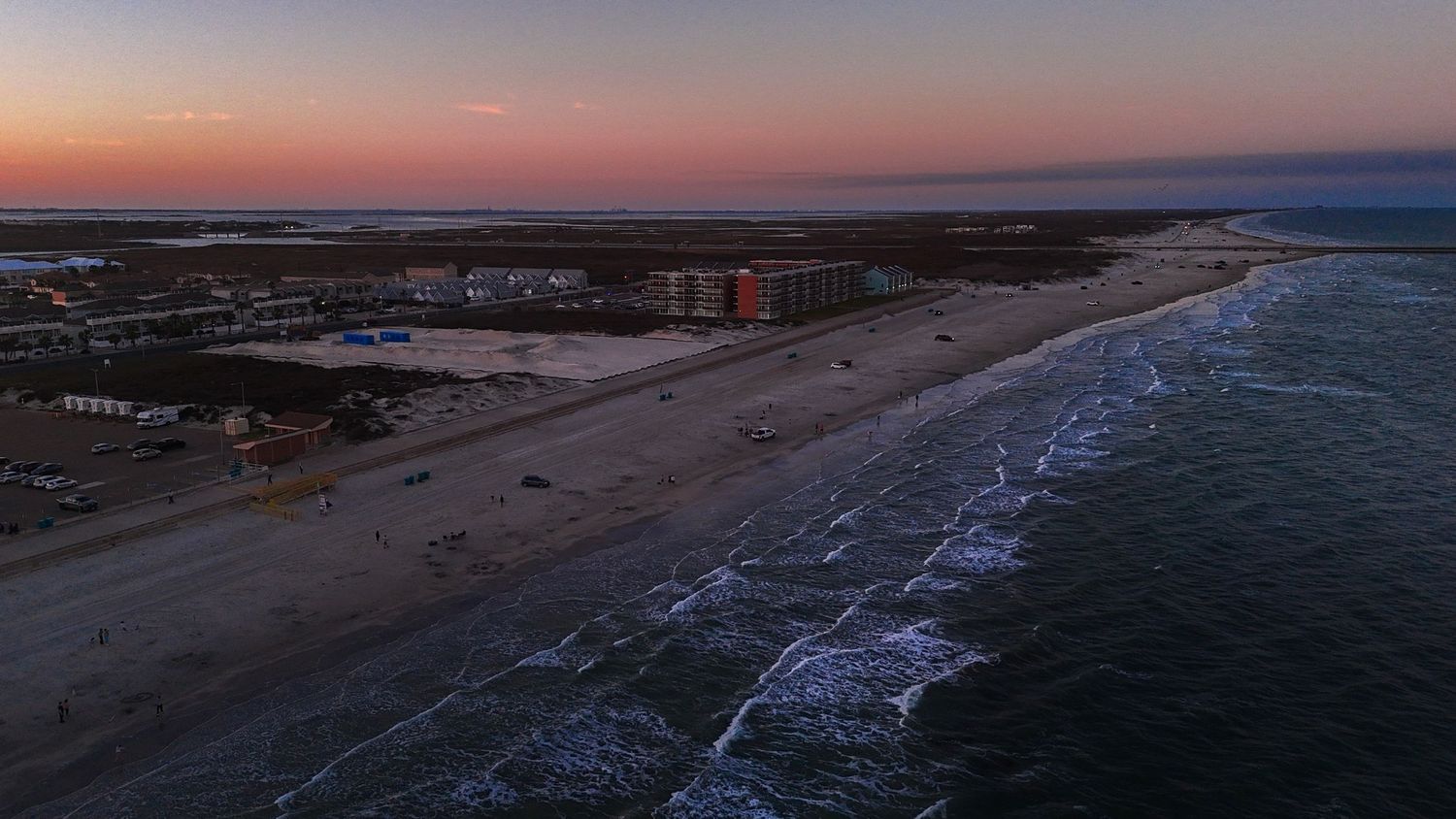 Aerial drone view of a beachfront at dusk with rolling waves and coastal buildings along the shoreline.