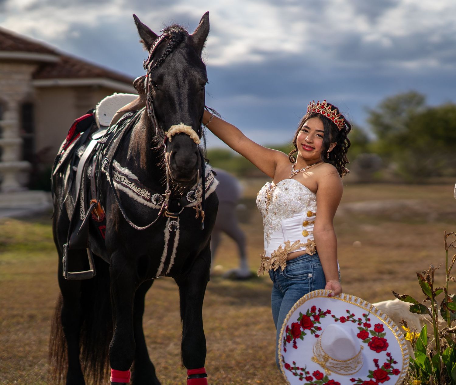 A woman in a white dress with red flowers poses playfully with a black horse in a rustic outdoor setting.
