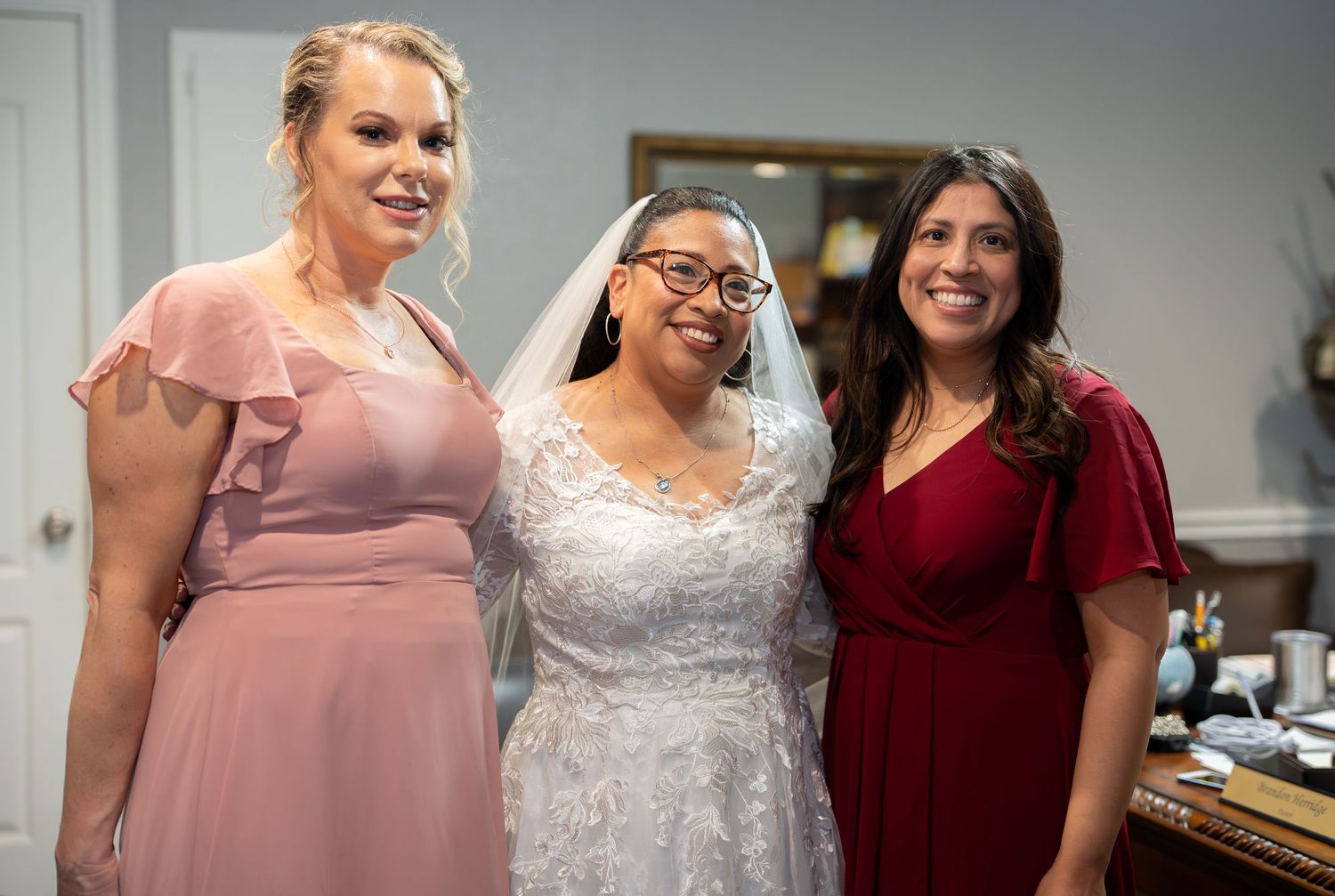 Bride in white lace gown poses with two smiling women in pink and red dresses at a wedding celebration.