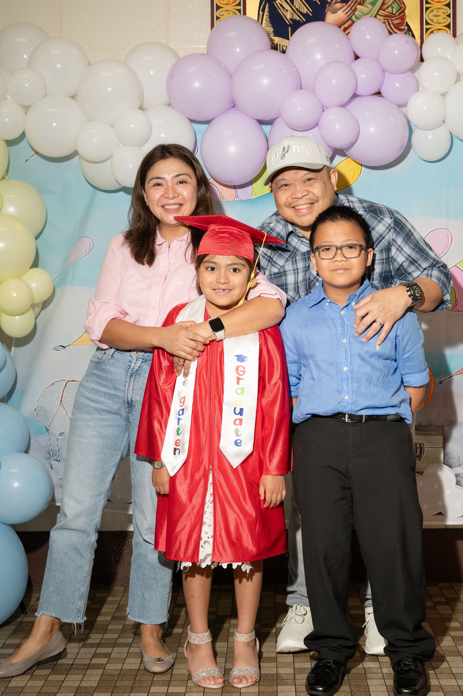 Family of four posing at a graduation ceremony, with a young girl in red cap and gown, surrounded by balloons.