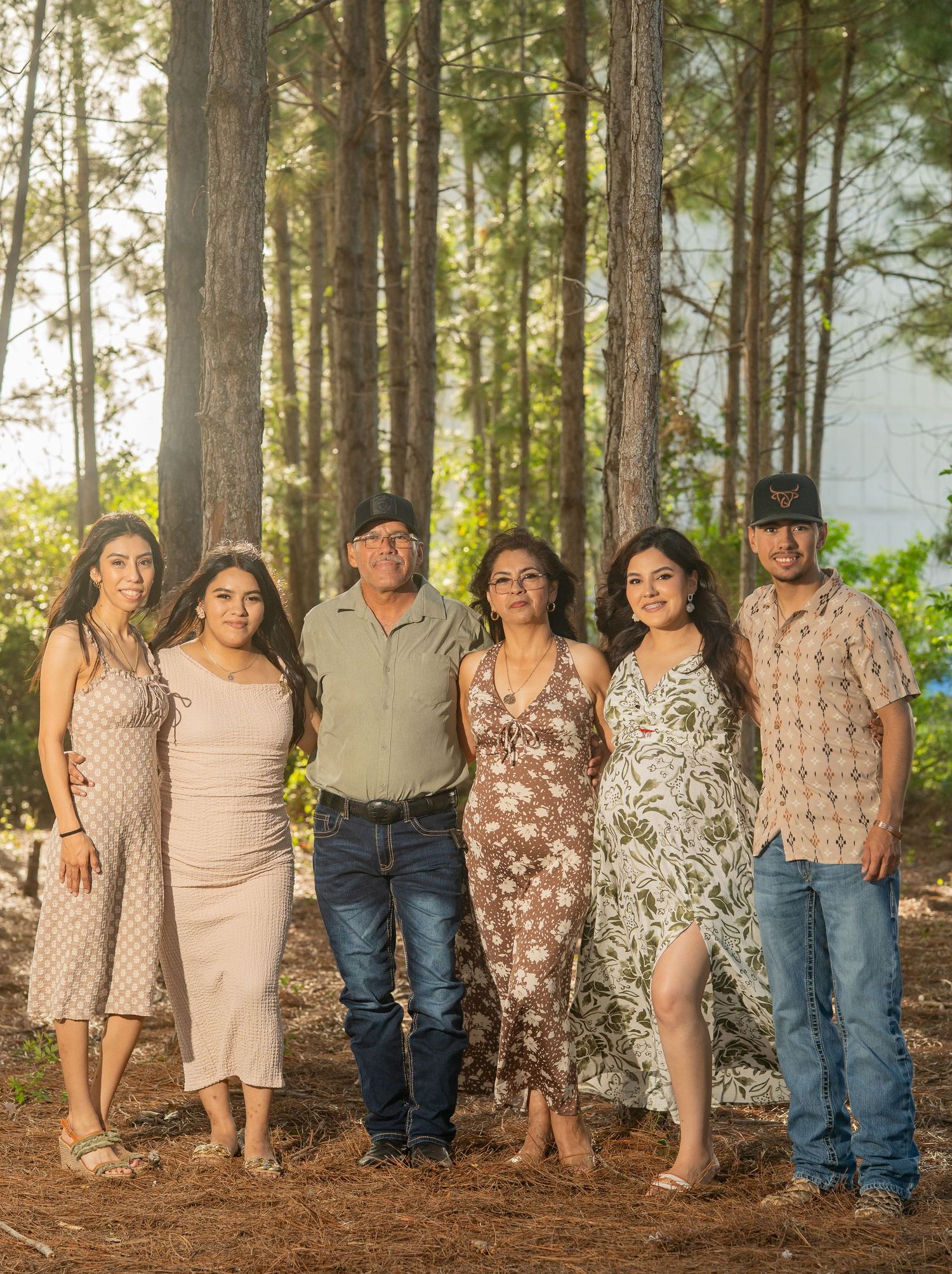 Family of six posing together in a pine forest, wearing coordinated outfits in warm tones for an outdoor portrait.